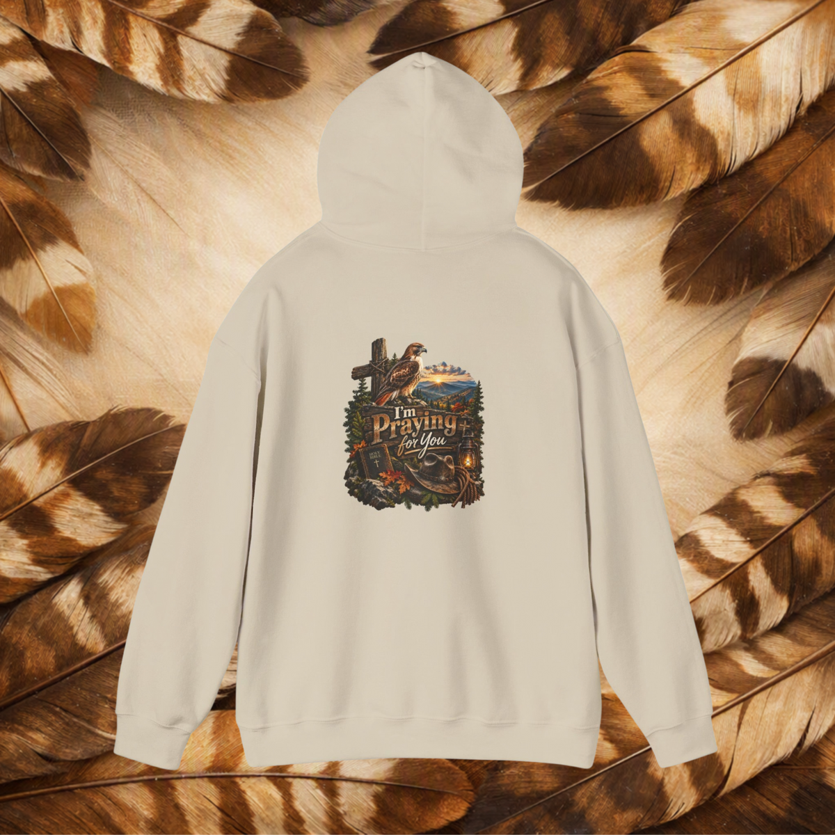 A cream-colored hoodie shown from the back, featuring a detailed graphic print with a cross, hawk, and the text 'I'm Praying For You'. The hoodie is laid flat on a background of brown and tan feathers.
