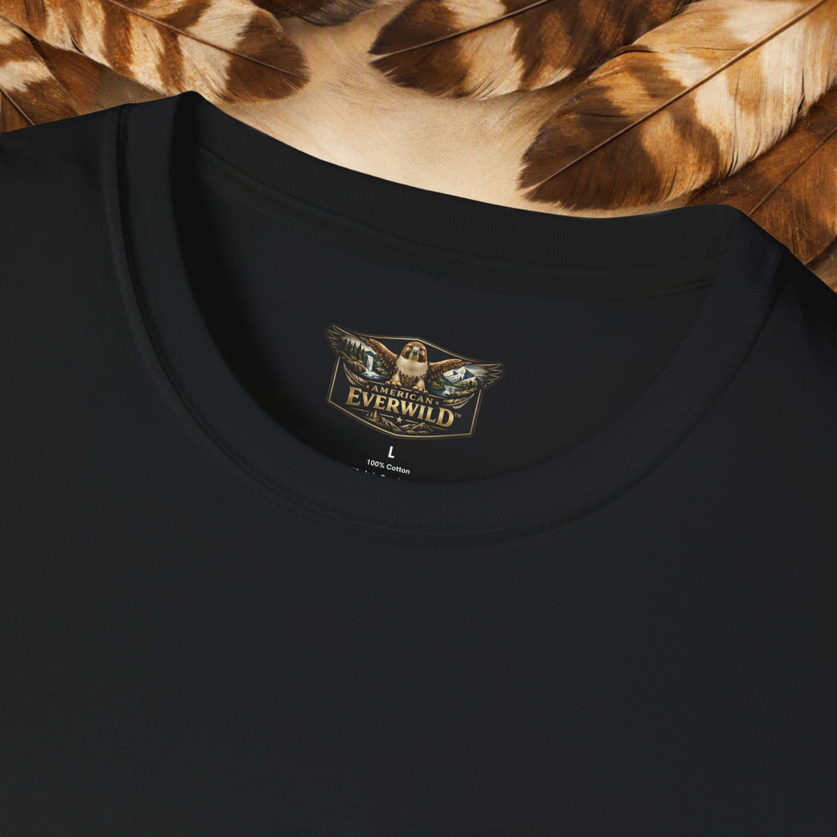 Close-up of a black crewneck t-shirt with a gold and brown "American Everwild" logo with a hawk with painted wings on the inside neck label, styled with brown and beige feathers at the top.