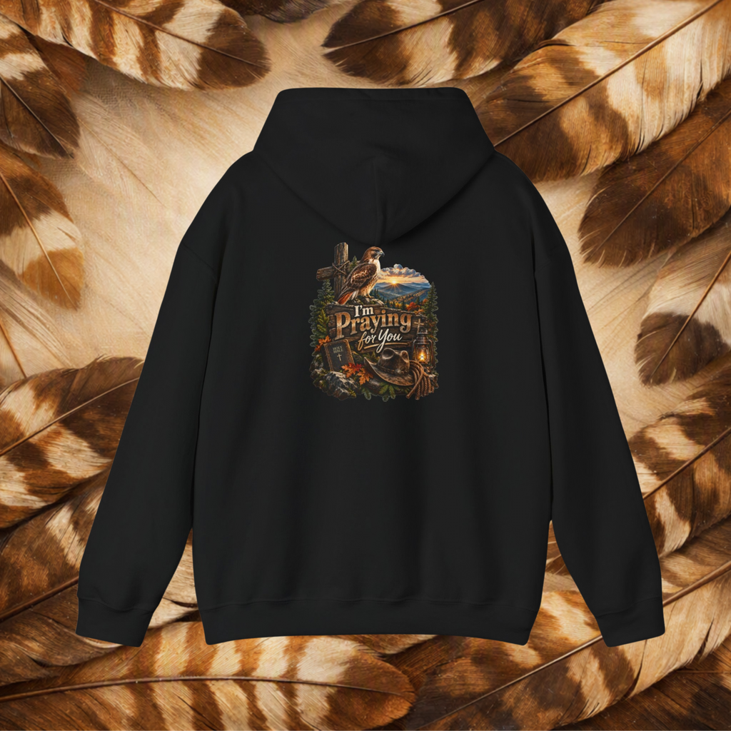 A black pullover hoodie shown from the back, featuring a detailed graphic print with a cross, a fishing rod, a cowboy hat, and the text 'I Am Praying For You' in a rustic outdoor theme. The hoodie is displayed flat against a background composed of brown and tan feathers.