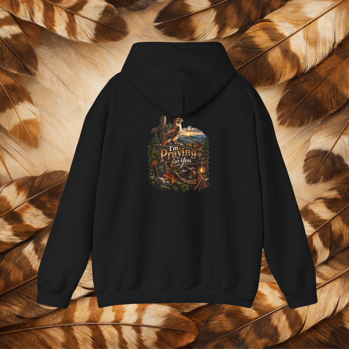 A black pullover hoodie shown from the back, featuring a detailed graphic print with a cross, a fishing rod, a cowboy hat, and the text 'I Am Praying For You' in a rustic outdoor theme. The hoodie is displayed flat against a background composed of brown and tan feathers.