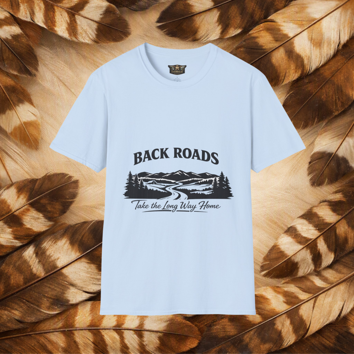 A light blue short-sleeve t-shirt with a black graphic print reading 'BACKROADS' and 'Take the Long Way Home' over a mountain and roadway scene, displayed flat against a background of brown and tan feathers.