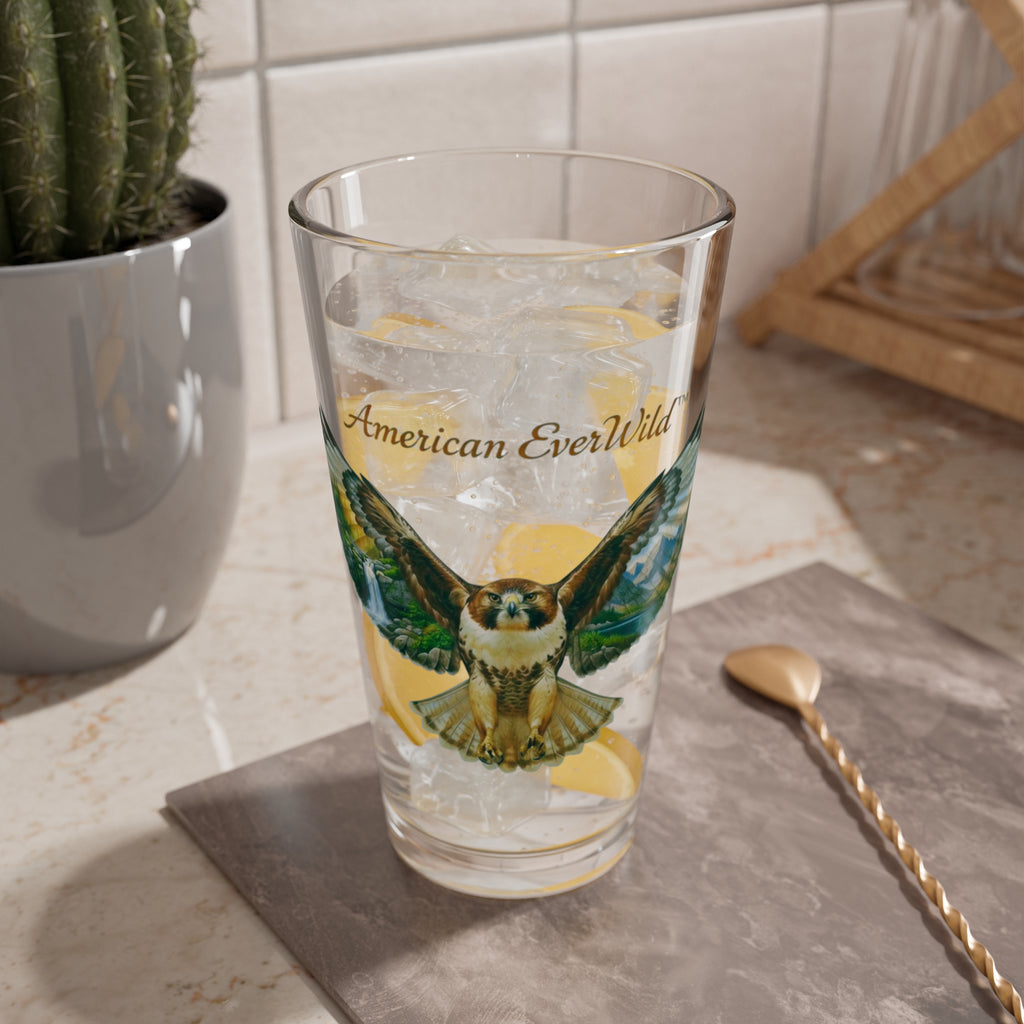 A clear drinking glass with a printed hawk illustration and the words 'American EverWild' on the front, shown filled with ice and lemon slices on a countertop in a kitchen setting.