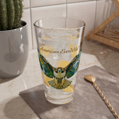 A clear drinking glass with a printed hawk illustration and the words 'American EverWild' on the front, shown filled with ice and lemon slices on a countertop in a kitchen setting.