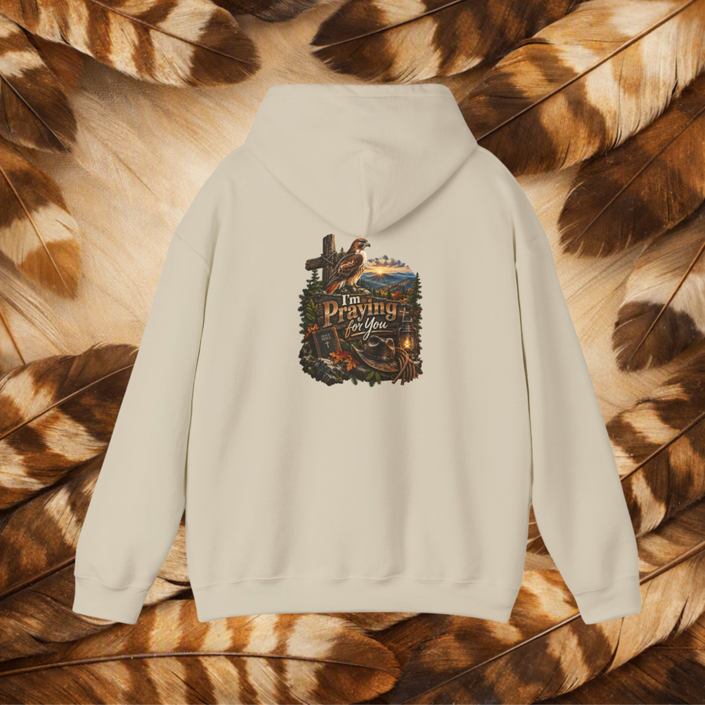An sand-colored hooded sweatshirt shown from the back, featuring a detailed graphic print with a cross, hawk, cowboy hat, and the text 'I'm Praying For You'. The hoodie is laid flat on a background of brown and black patterned feathers.