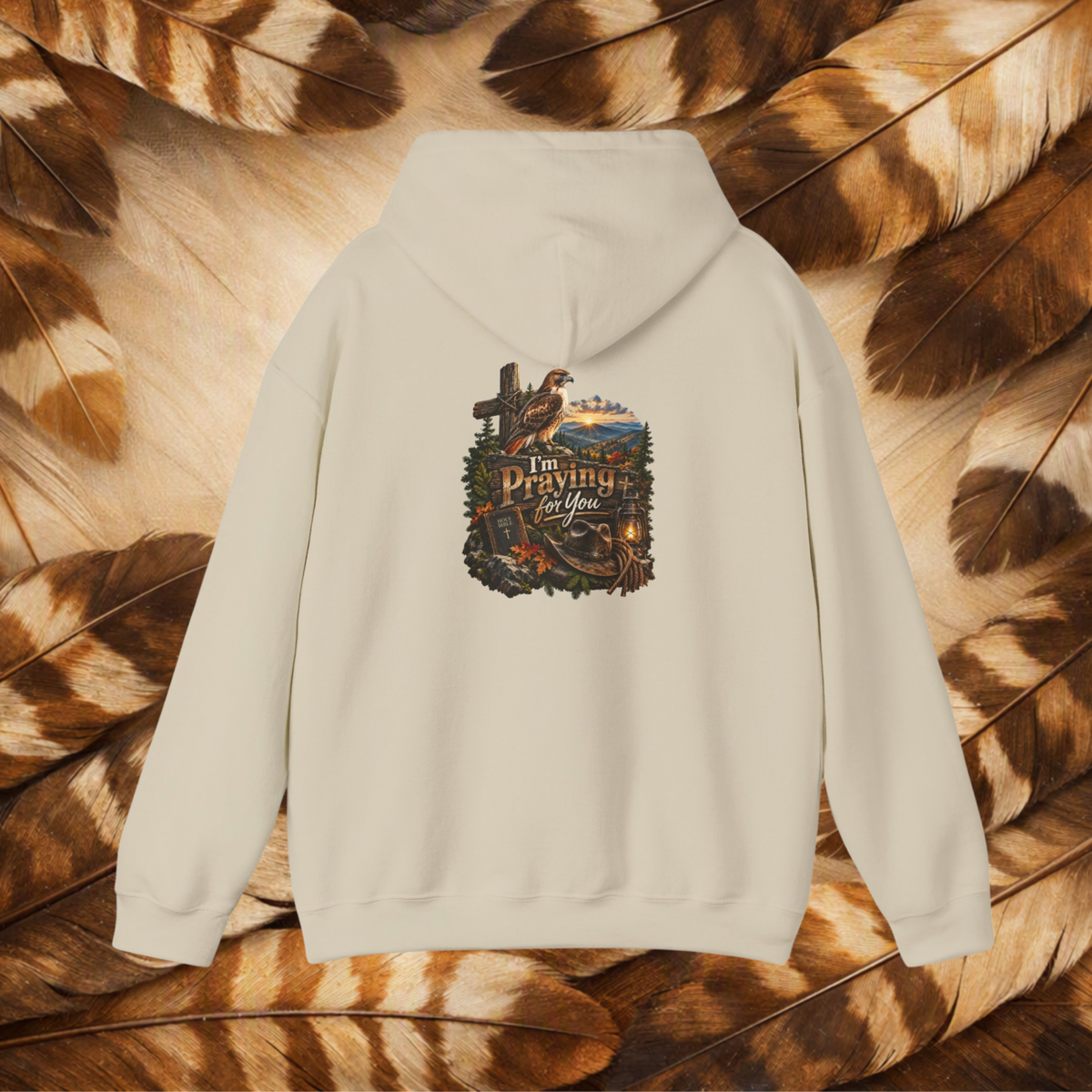 An sand-colored hooded sweatshirt shown from the back, featuring a detailed graphic print with a cross, hawk, cowboy hat, and the text 'I'm Praying For You'. The hoodie is laid flat on a background of brown and black patterned feathers.