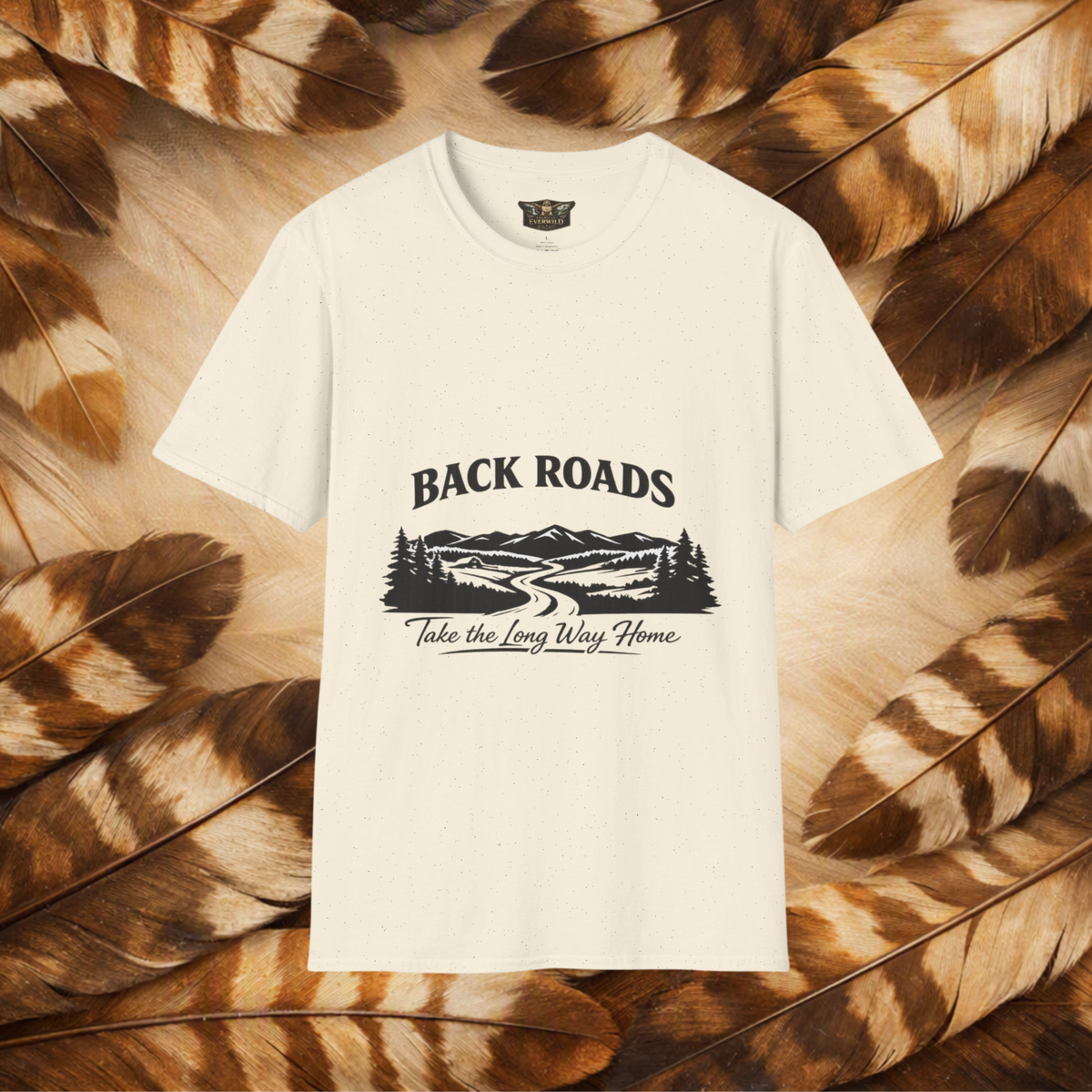 An off-white short-sleeve t-shirt with a black graphic print reading 'BACKROADS' and 'Take the Long Way Home' over a mountain and roadway illustration, displayed flat against a background of brown and beige feathers.