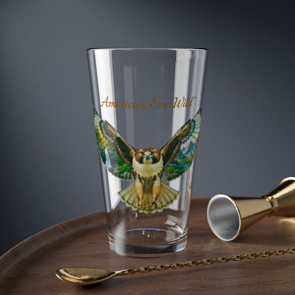 A clear drinking glass featuring a printed illustration of a red tailed hawk bird of prey with painted wings and the text 'American EverWild' in gold, shown on a wooden surface with gold-toned bar tools against a dark gray background.