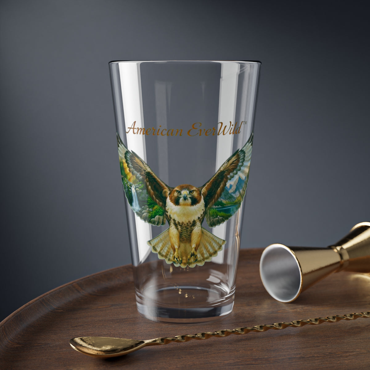 A clear drinking glass featuring a printed illustration of a red tailed hawk bird of prey with painted wings and the text 'American EverWild' in gold, shown on a wooden surface with gold-toned bar tools against a dark gray background.