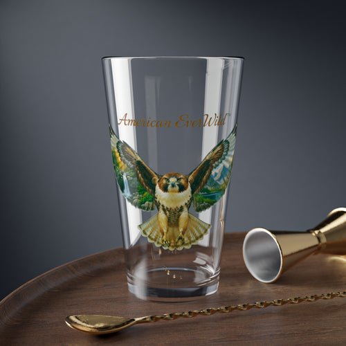 A clear drinking glass featuring a printed illustration of a red tailed hawk bird of prey with painted wings and the text 'American EverWild' in gold, shown on a wooden surface with gold-toned bar tools against a dark gray background.