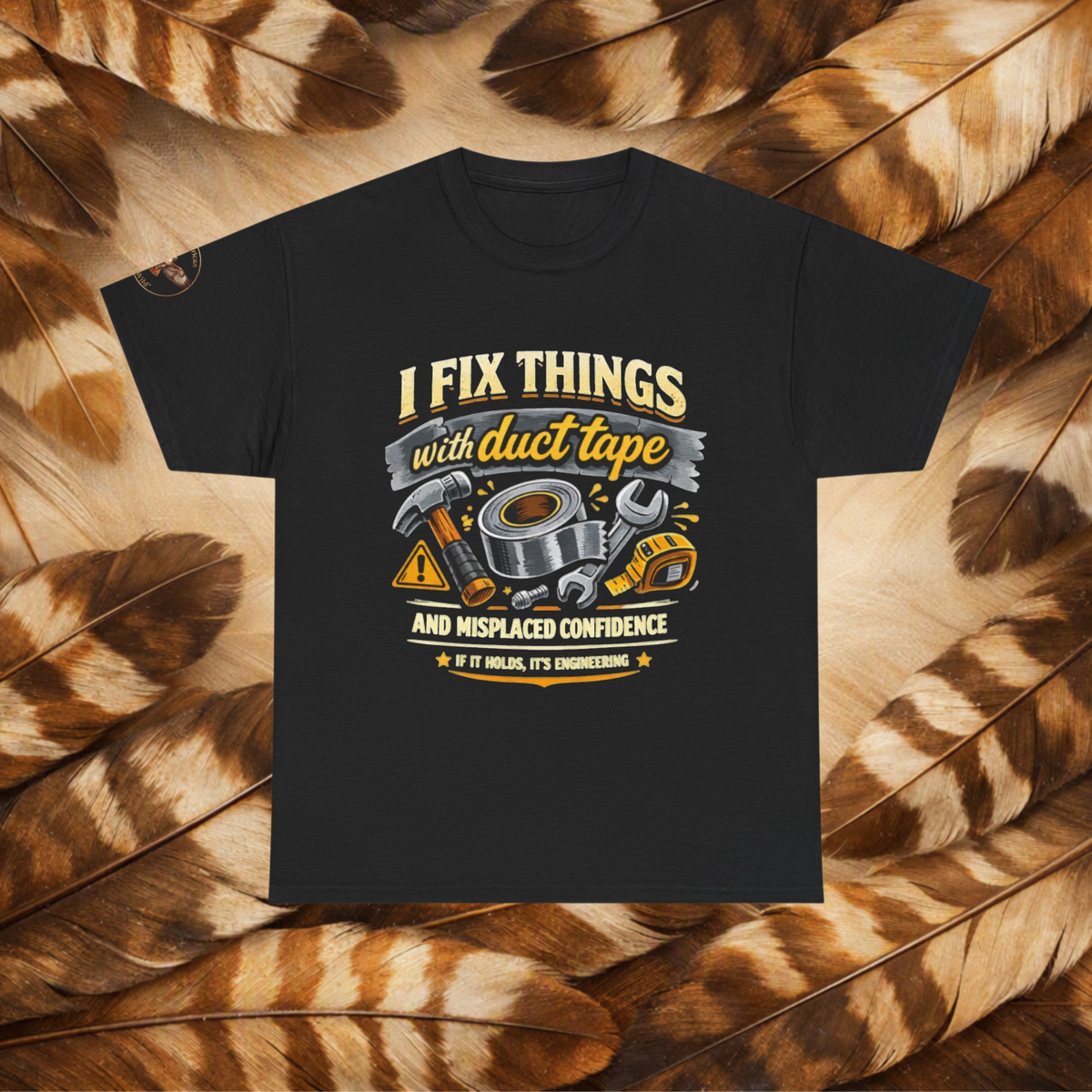 Black t-shirt with a front view graphic with tools and text that reads'I Fix Things With Duct Tape and Misplaced Confidence, If it Holds It's Engineering', laid flat on a background of cream and brown feathers.