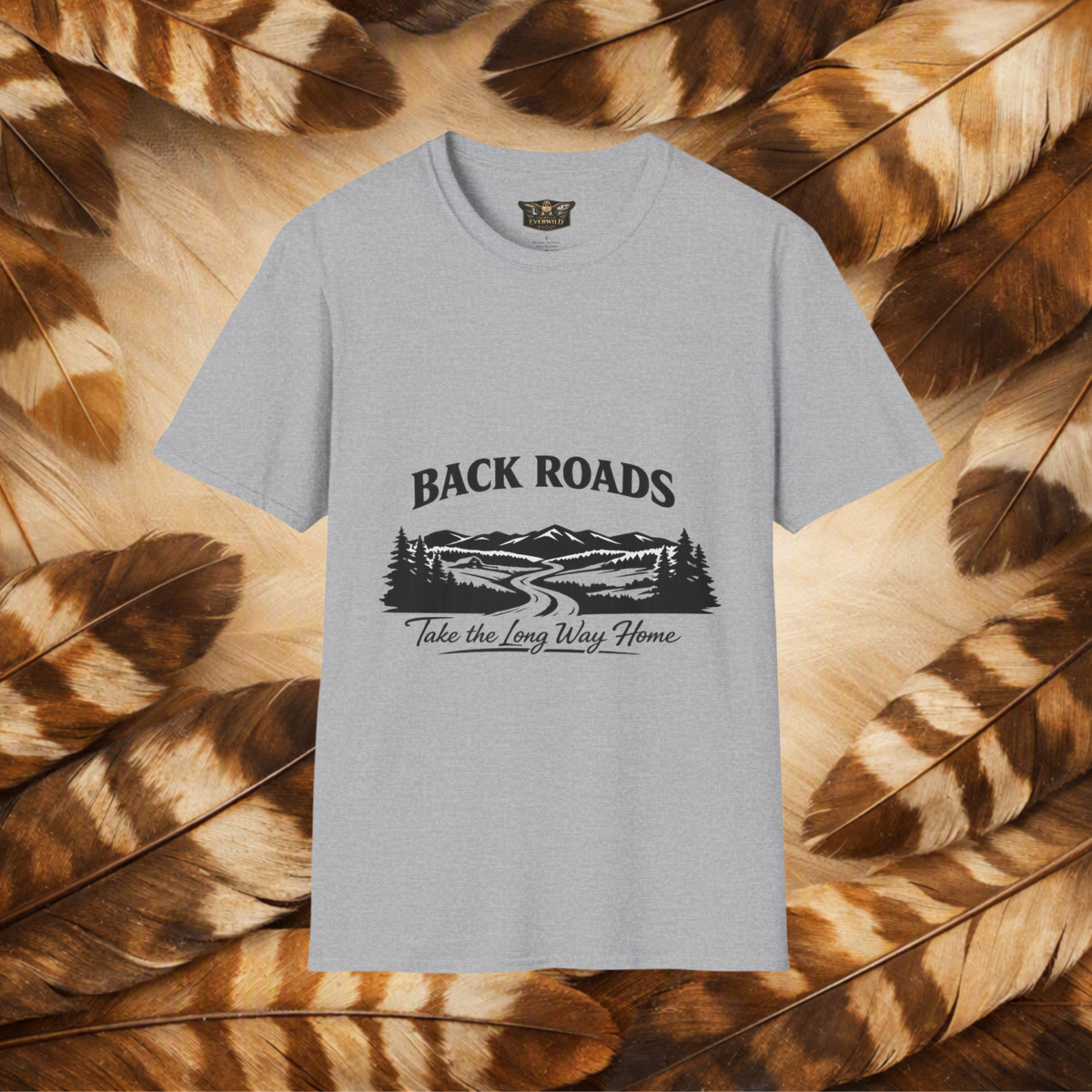 A light gray crewneck t-shirt with a black graphic print reading 'BACKROADS Take the Long Way Home' above a mountain and roadway scene, displayed flat against a background of brown and tan feathers.