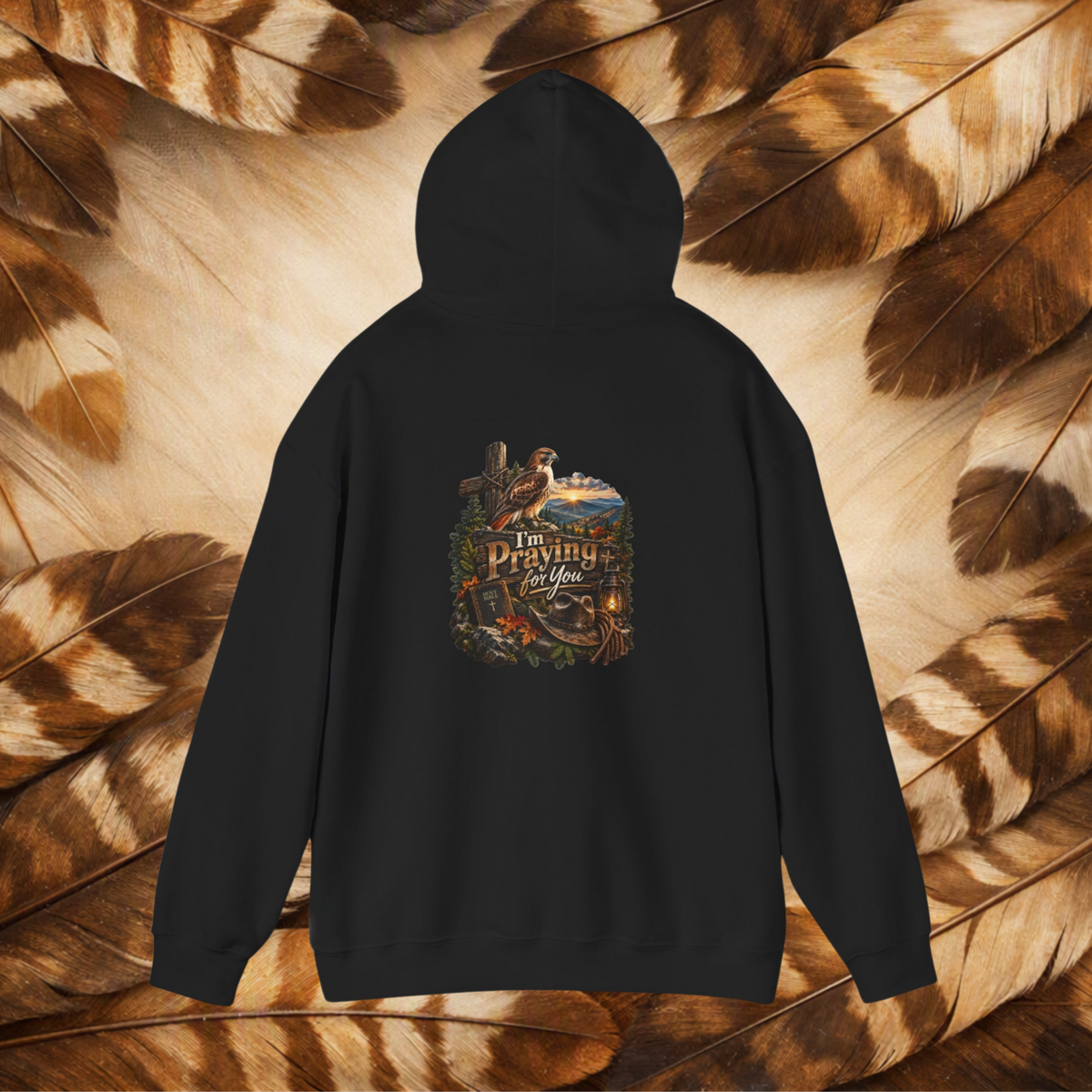 A black pullover hoodie shown from the back, featuring a large graphic print with a hawk, mountain, and the text 'I'm Praying For You.' The hoodie is laid flat on a background of brown and tan feathers.