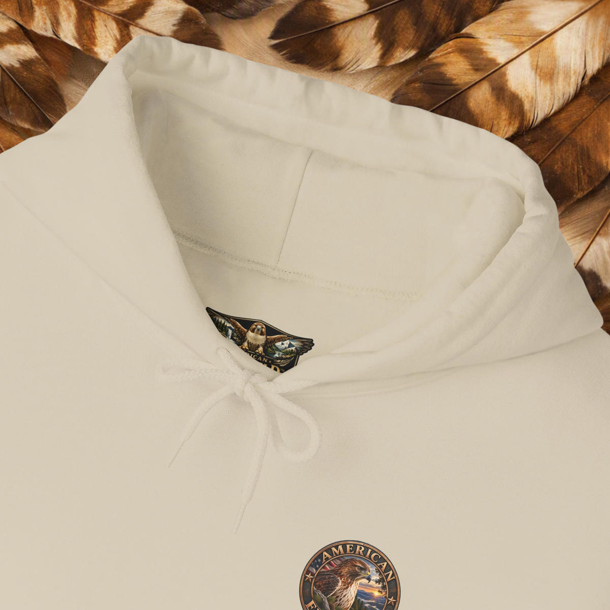 An sand-colored hooded sweatshirt laid flat on a background of brown and beige feathers. The hoodie features a drawstring hood, a small embroidered patch on the chest, and a brand tag inside the collar.