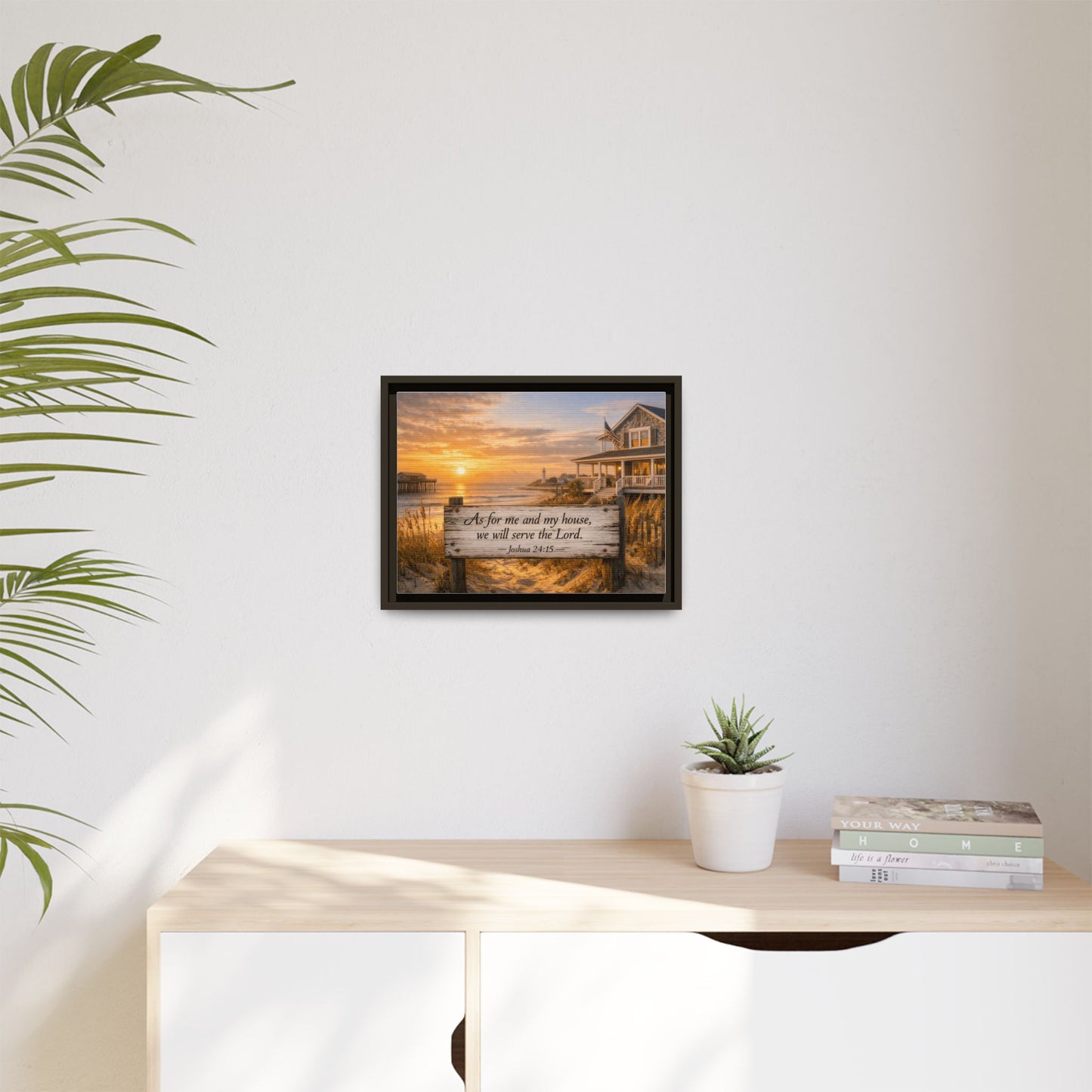Small framed coastal scripture wall art on white wall with plant pot and stacked books.