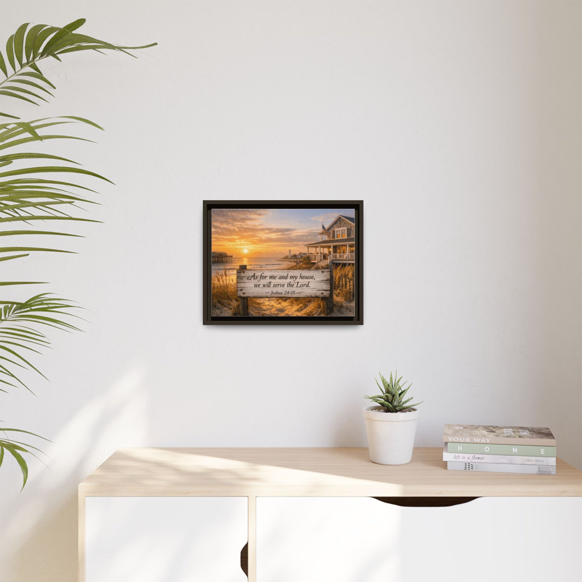 Small framed coastal scripture wall art on white wall with plant pot and stacked books.
