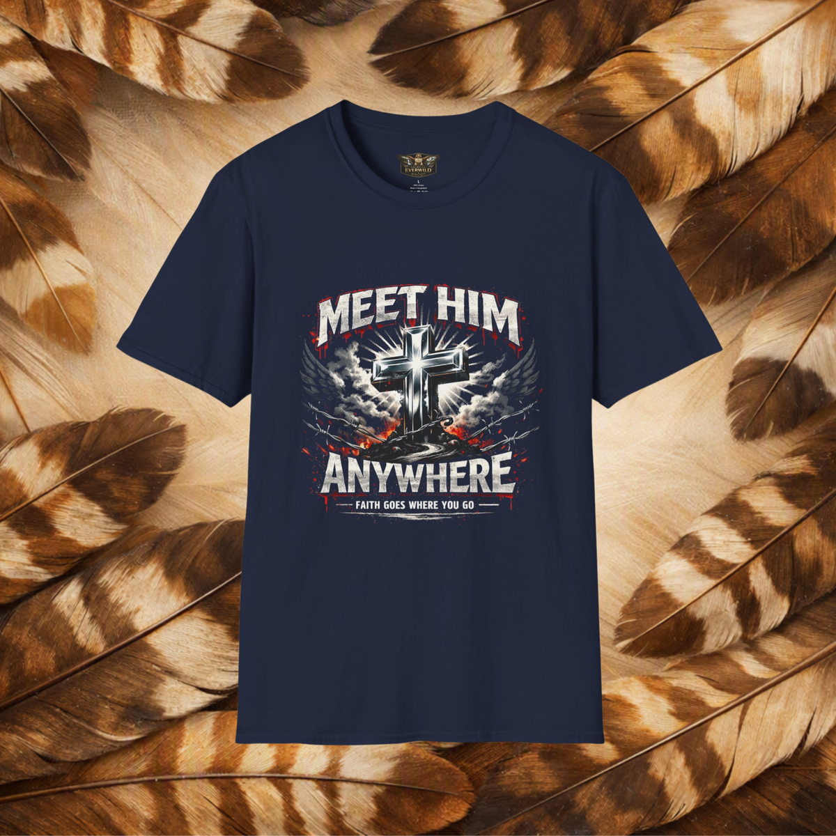 A navy short-sleeve graphic t-shirt displayed flat against a background of brown and beige feathers. The shirt features a large front graphic with a metallic silver cross, white and red text reading 'MEET HIM ANYWHERE', and a banner that says 'FAITH GOES WHERE YOU GO'.