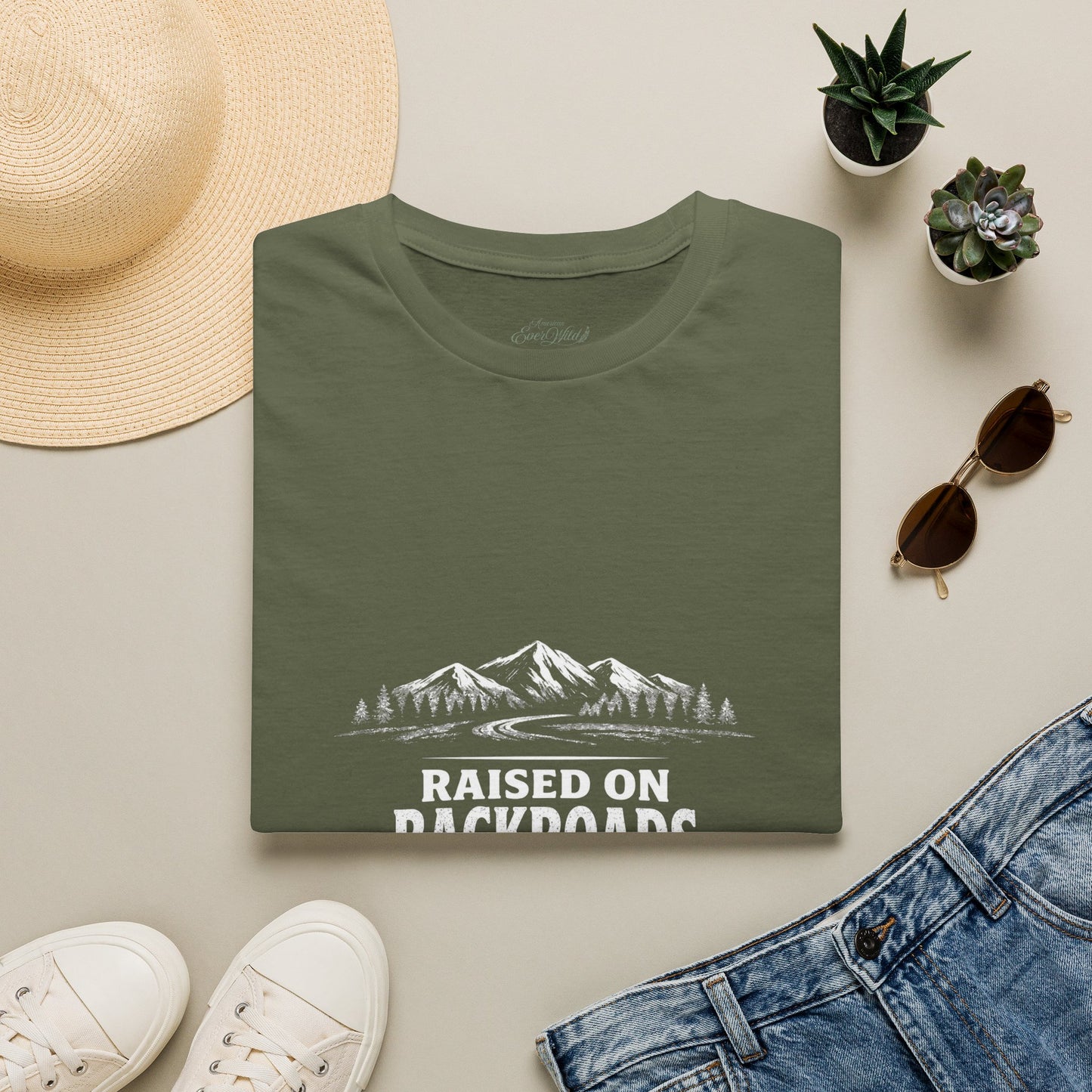 Military green crewneck T-shirt folded flat with mountain landscape “Raised on Backroads” graphic.