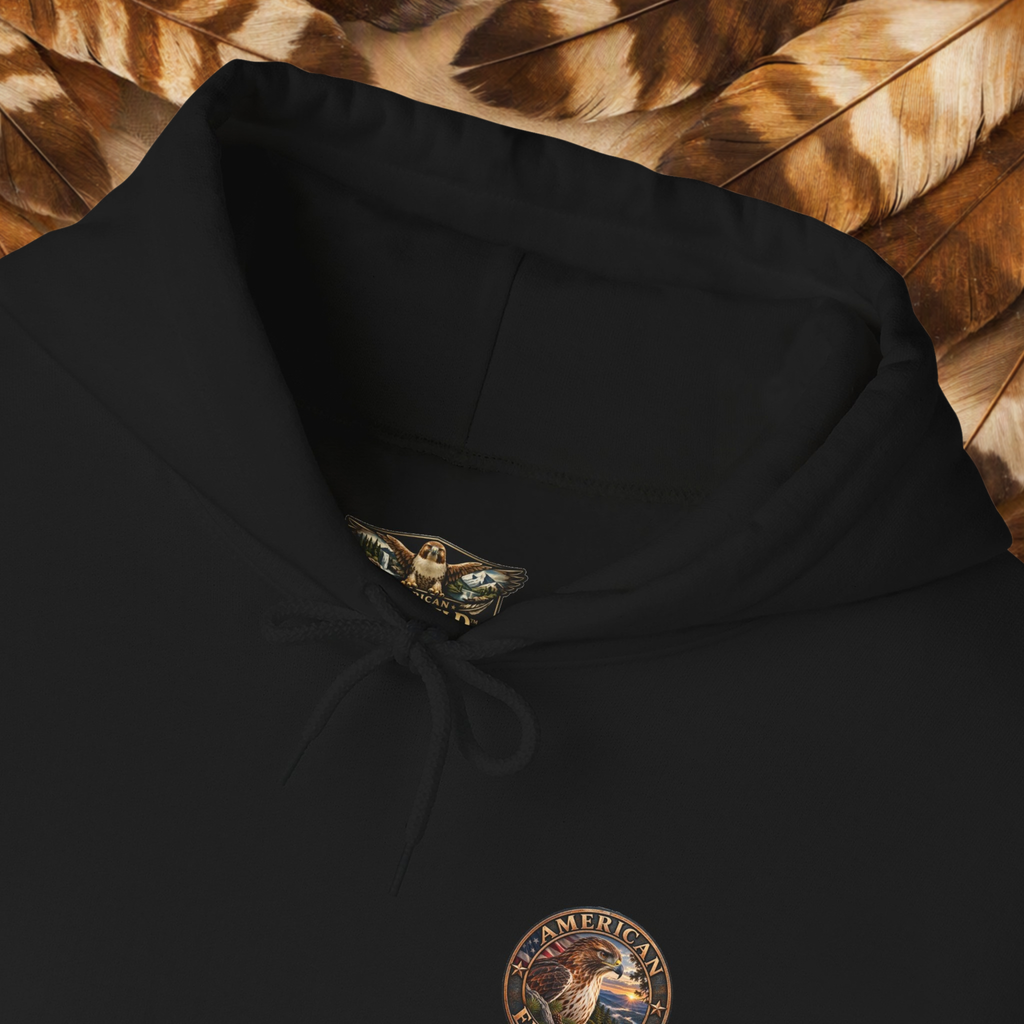 Close-up of a black pullover hoodie laid flat on a bed of brown feathers, showing the drawstring hood, a small embroidered chest graphic, and a branded neck label.