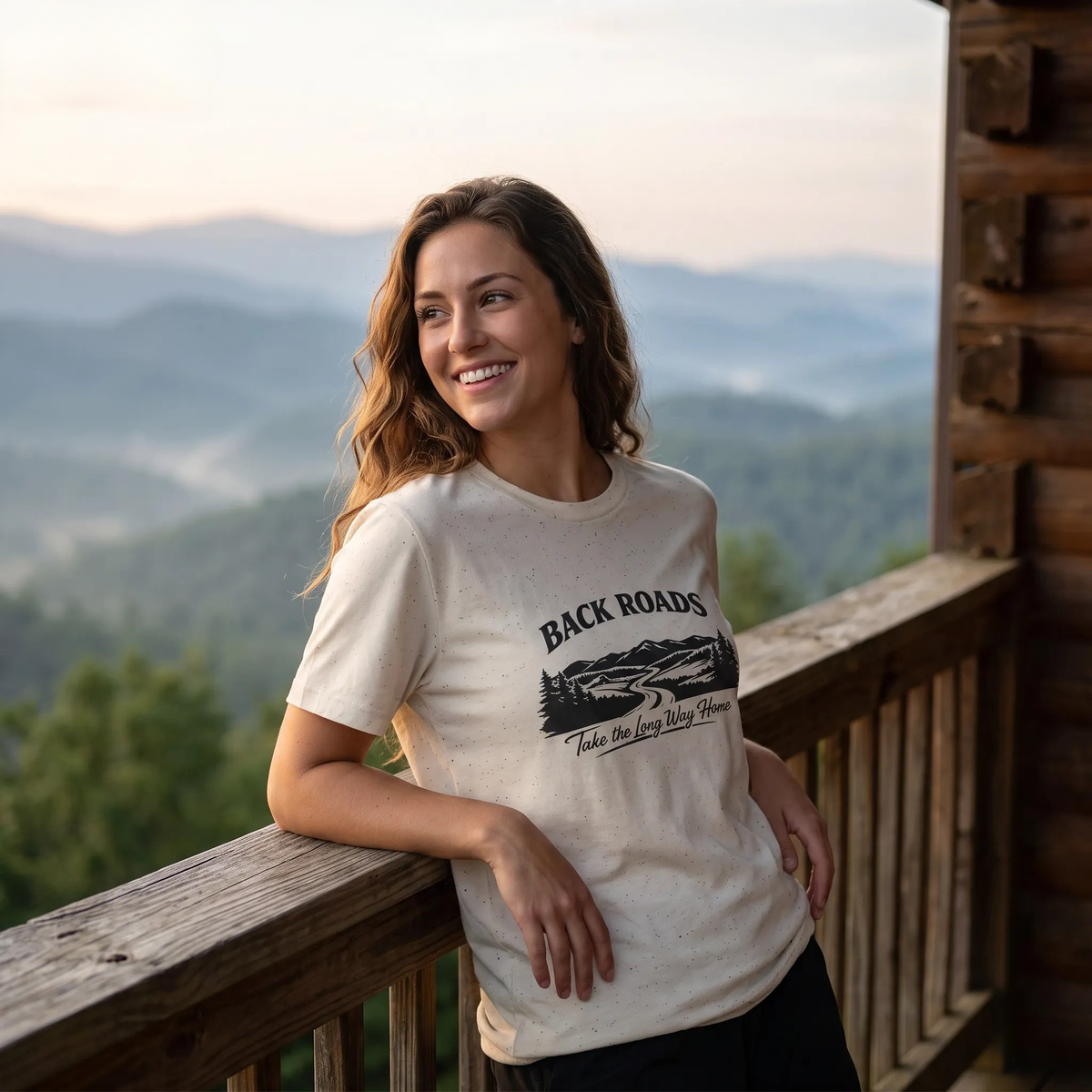 A woman wearing a cream-colored graphic t-shirt with 'BACKROADS' and a mountain road illustration with "Take The Long Way Home" below, she is leaning on a wooden balcony railing with a misty mountain landscape in the background.