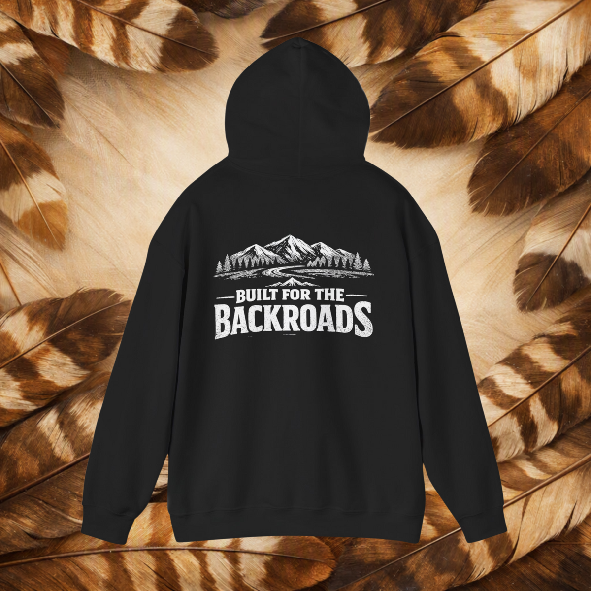 A black hoodie shown from the back, featuring a white graphic of mountains and trees with the text 'BUILT FOR THE BACKROADS'. The hoodie is displayed flat against a background of brown and beige feathers.