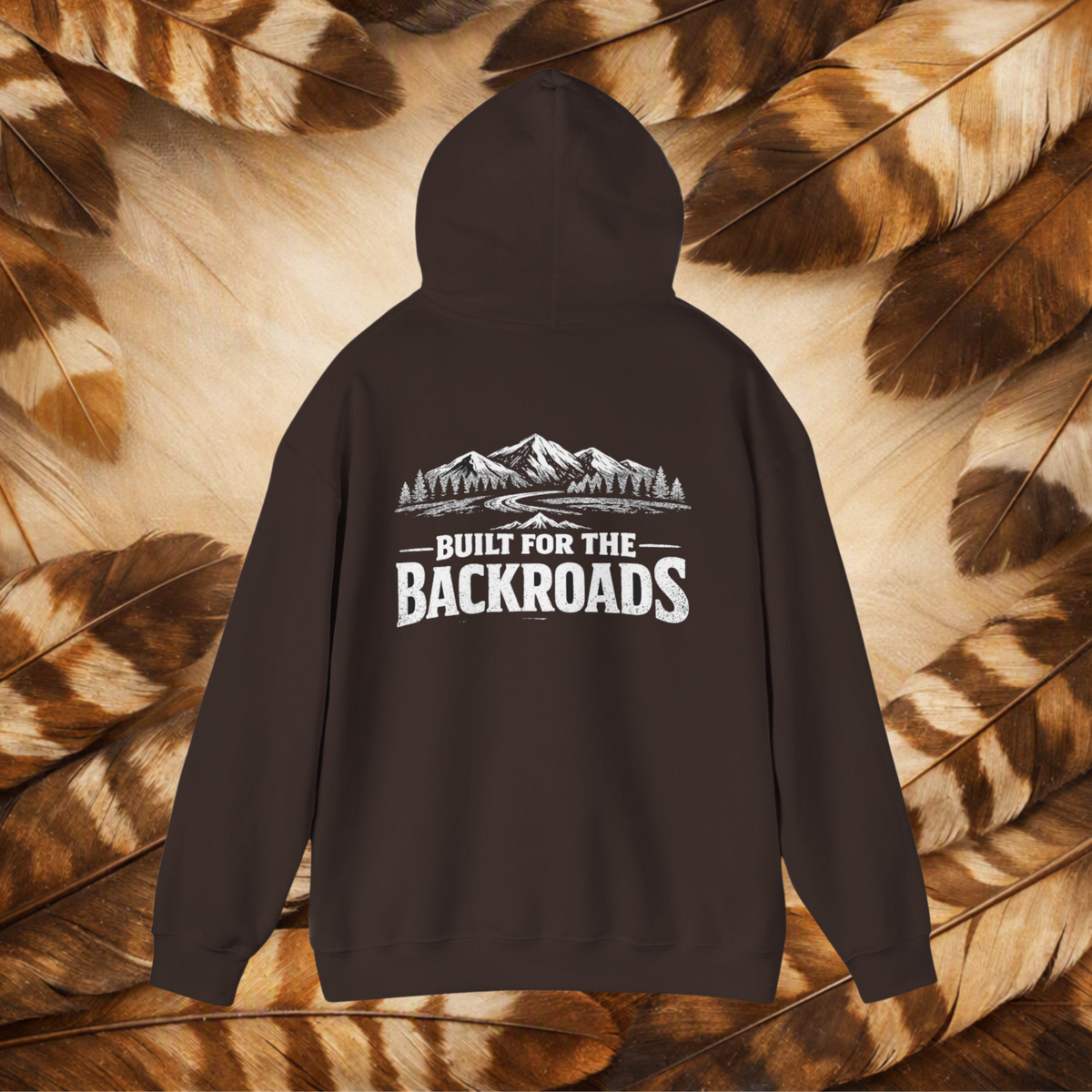 A chocolate brown hoodie shown from the back, featuring a white graphic of mountains and trees with the text 'BUILT FOR THE BACKROADS'. The hoodie is displayed flat against a background of brown and beige feathers.