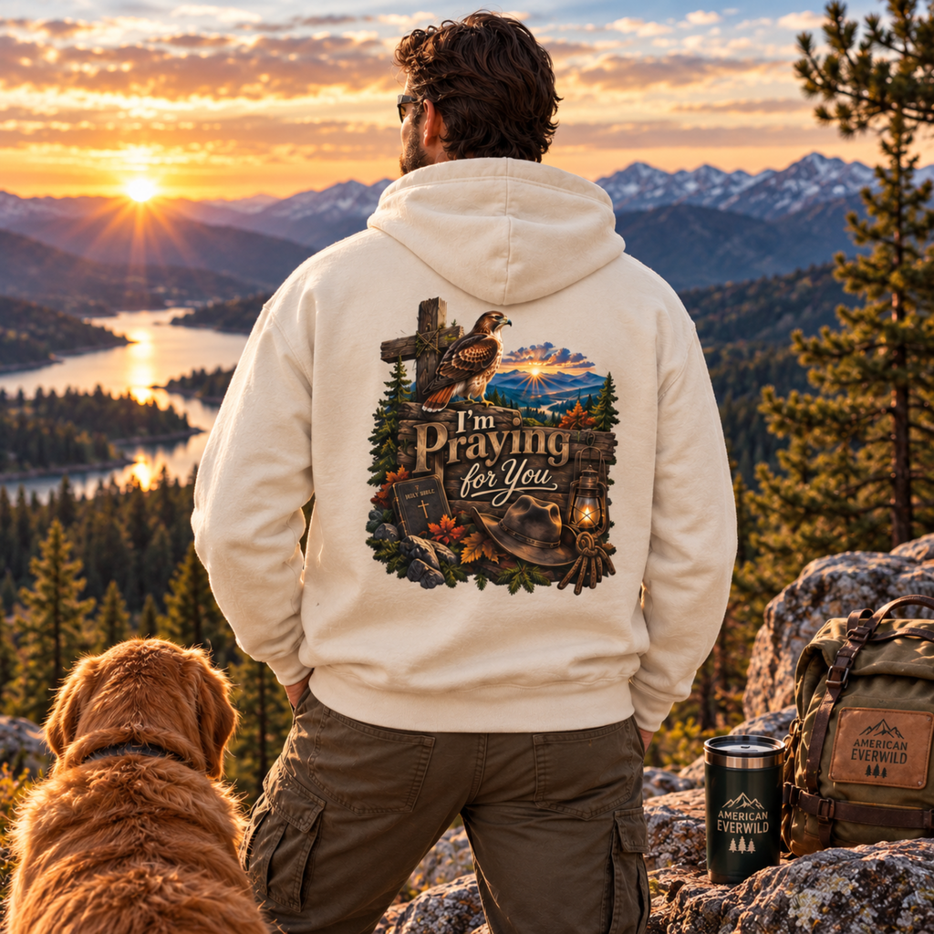 A man stands on a mountain overlook at sunset with a lake and forest in the background, wearing a cream hoodie with a large back graphic that reads “I’m Praying For You” with a cross, bird, mountains, and lantern. A dog sits at his left, and a backpack and tumbler are at his right.