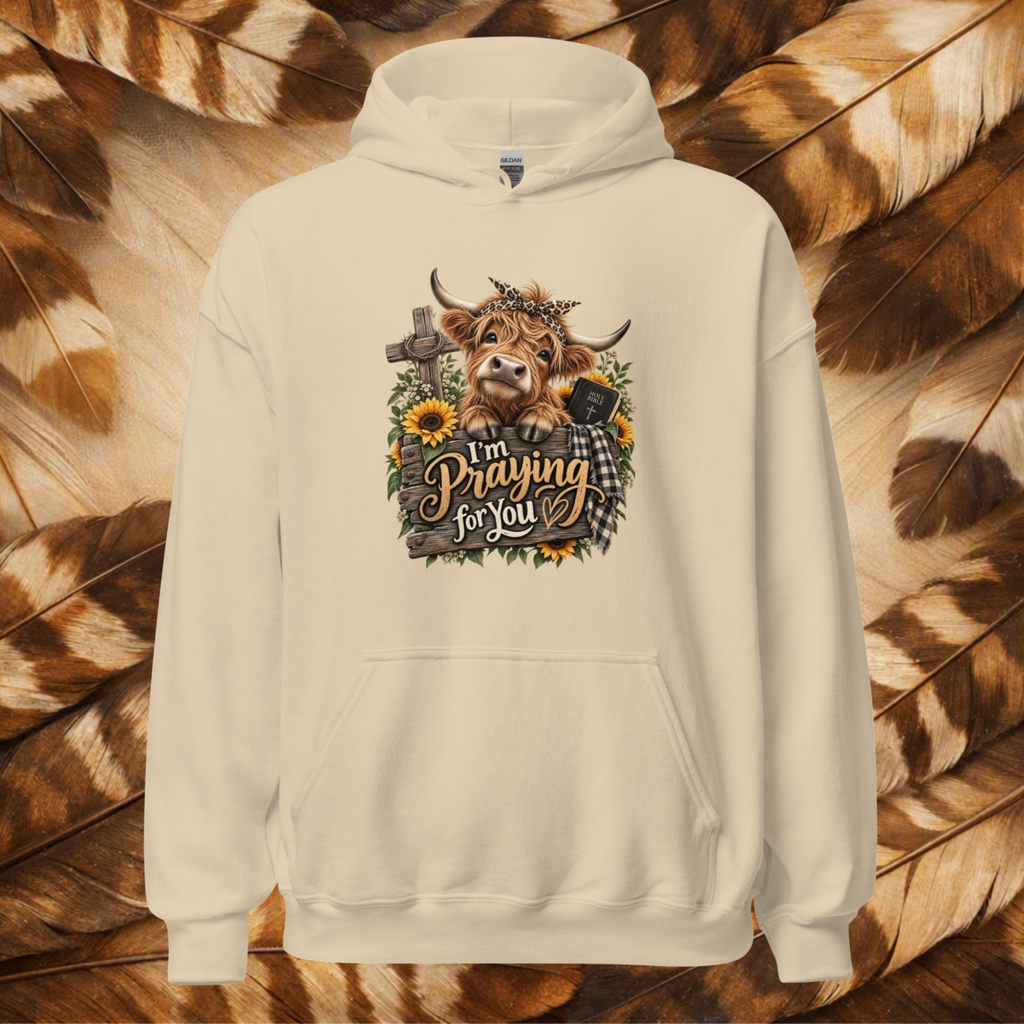 Sand-colored front-lay hoodie with a cute country cow leaning over a fence that says "I'm Praying For You." Has sunflowers and wild flowers with a cross and bible on a background a cream and brown feathers.