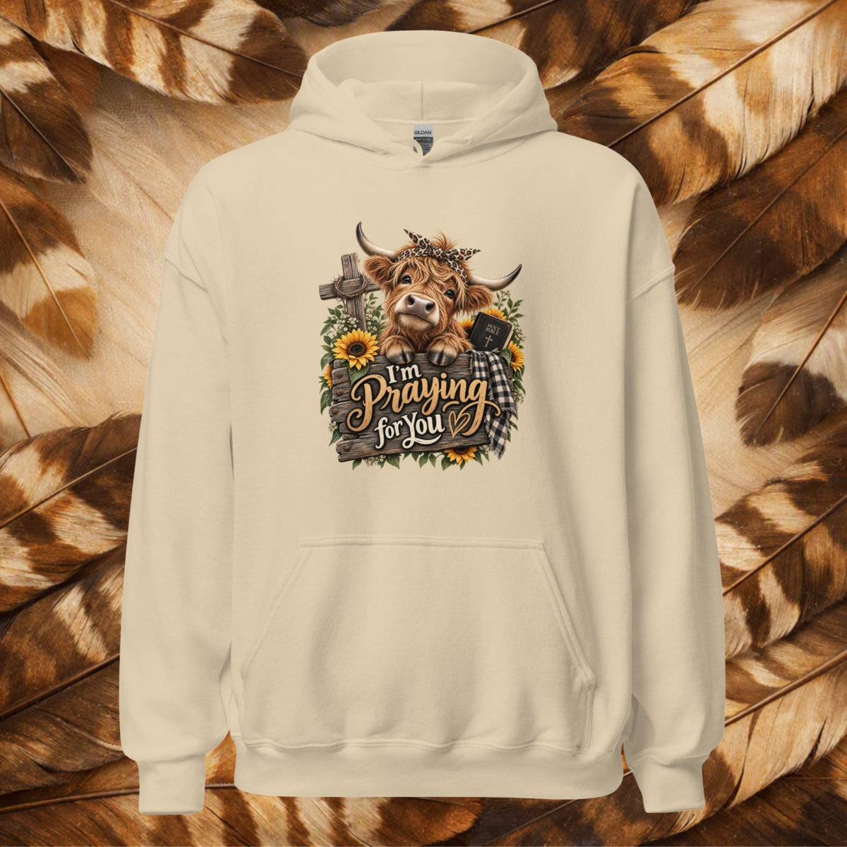 Sand-colored front-lay hoodie with a cute country cow leaning over a fence that says "I'm Praying For You." Has sunflowers and wild flowers with a cross and bible on a background a cream and brown feathers.