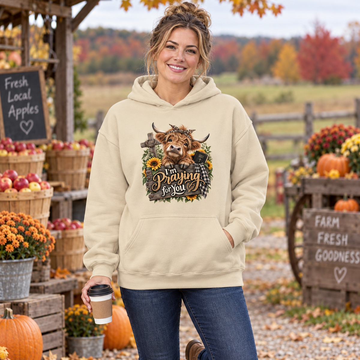 Woman at farmers market with coffee cup. The background has fall color flowers with fruits and pumpkins while she wears a sand-colored hoodie with a playful cute country cow with a bow that is leaning across a fence with the text "I'm Praying for You" on the front chest.