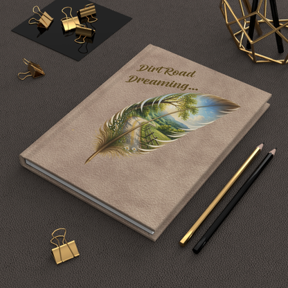 A beige hardcover notebook or journal lying on a dark textured surface, decorated with the words 'Dirt Road Dreaming.