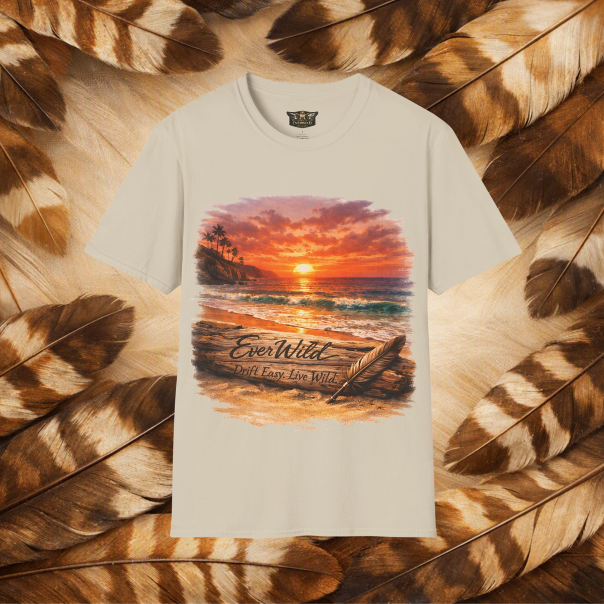 A sand color tee with a graphi of a sunset over the beach and a drift log with carved text 'EverWild—Drift Easy-Live wild' and feather quill leaning on the log in the sand. Flat lay on a background of tan and brown feathers.