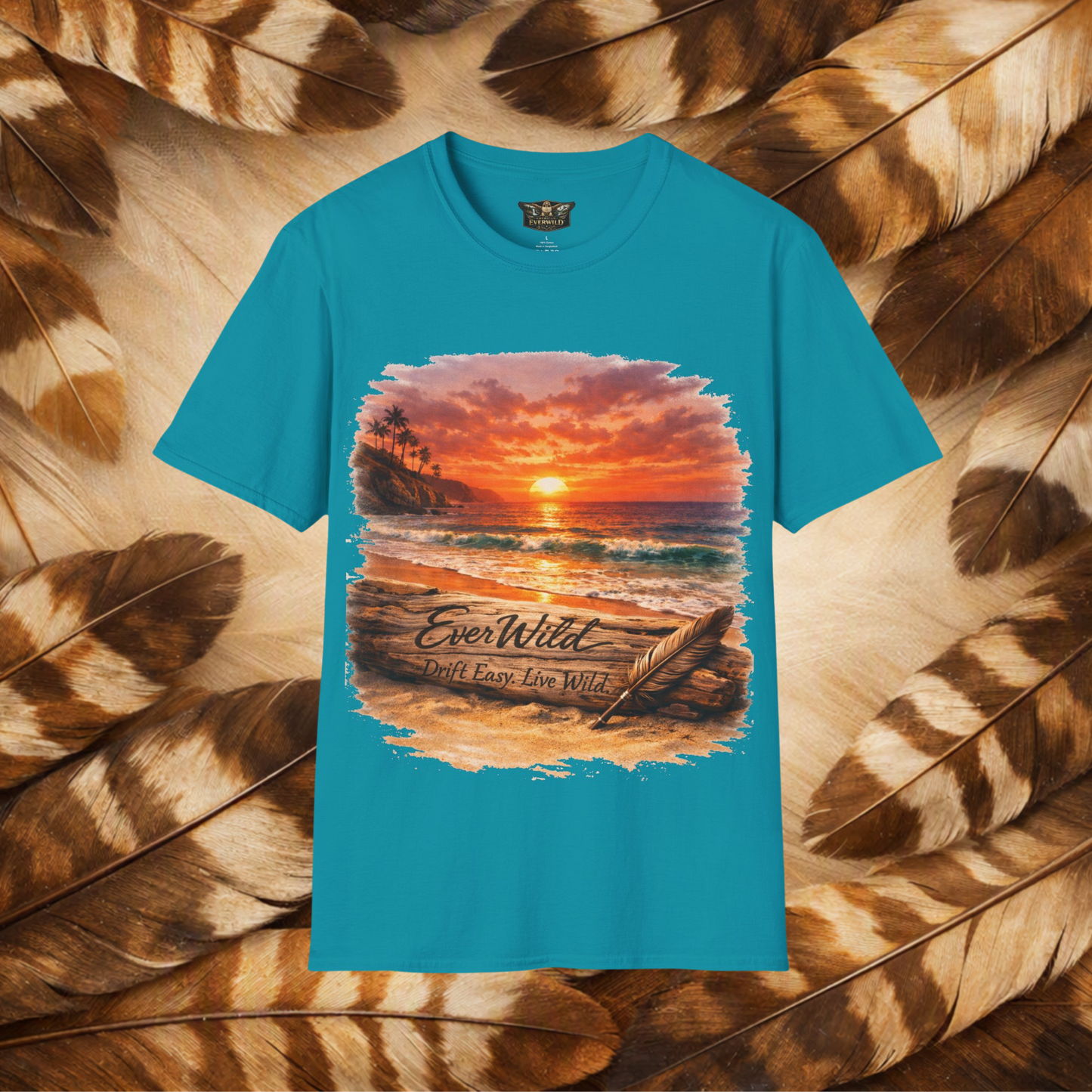 A tropical blue short-sleeve t-shirt with a large rectangular graphic print of a beach sunset scene. The graphic features the text 'EverWild Drift Easy- Live Wild' laid flat on a background of cream and brown feathers.