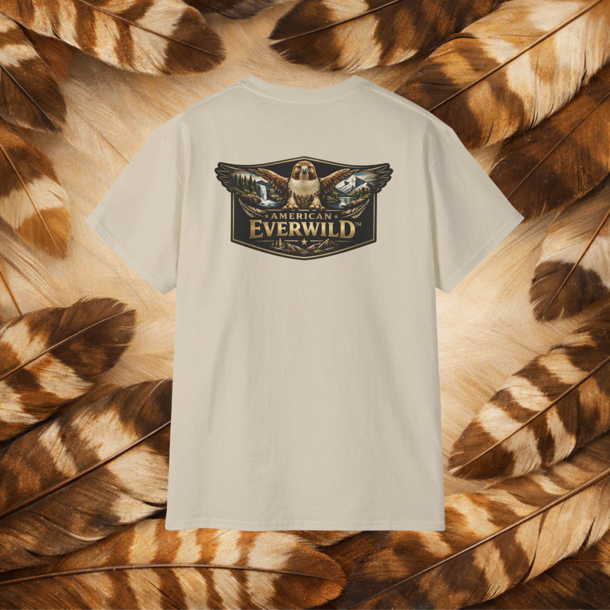 A sand-colored short-sleeve t-shirt with a large graphic on the back featuring a hawk and the text 'AMERICAN EVERWILD'. The shirt is displayed flat against a background of brown and beige feathers.