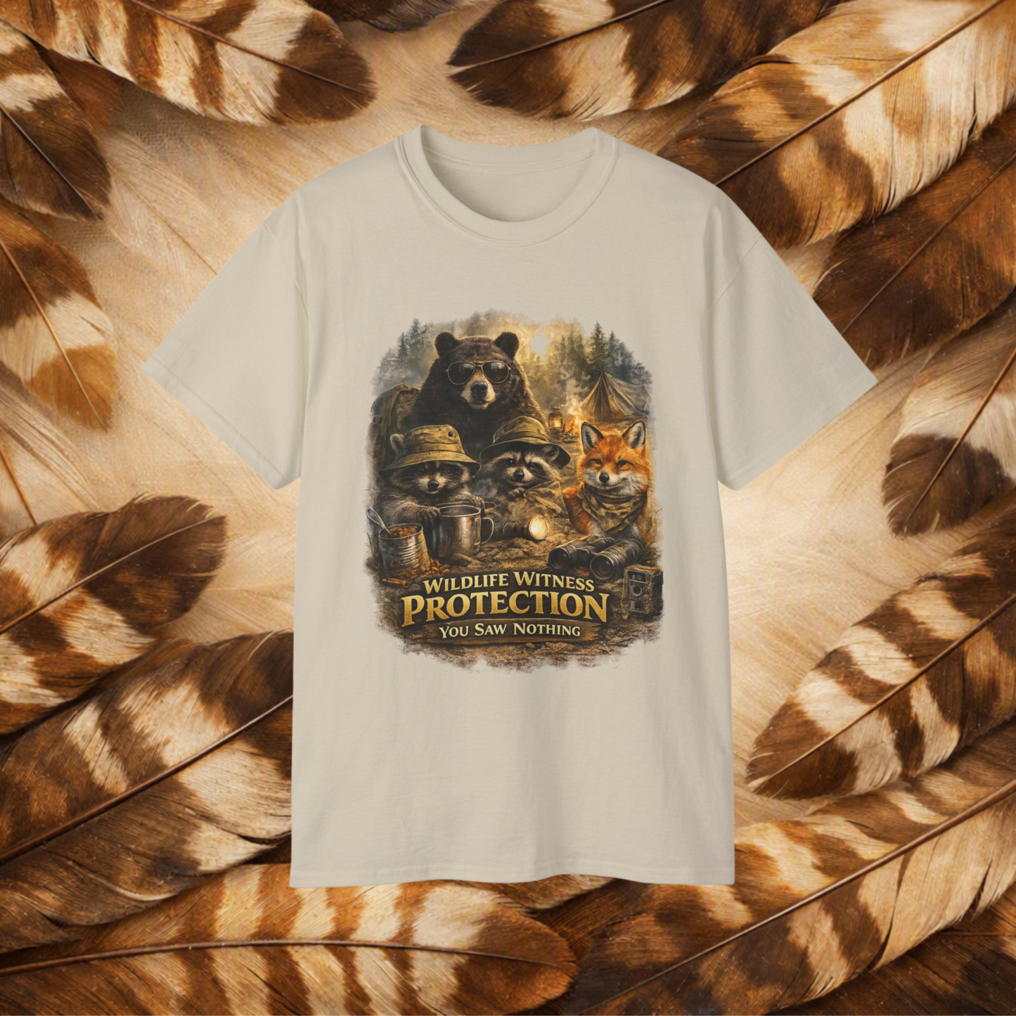 A Sand-colored short-sleeve t-shirt with a central graphic of raccoons, a bear, and a fox wearing hats or sunglasses, with the text 'WILDLIFE WITNESS PROTECTION YOU SAW NOTHING'. The shirt is displayed flat against a background of brown and beige feathers.