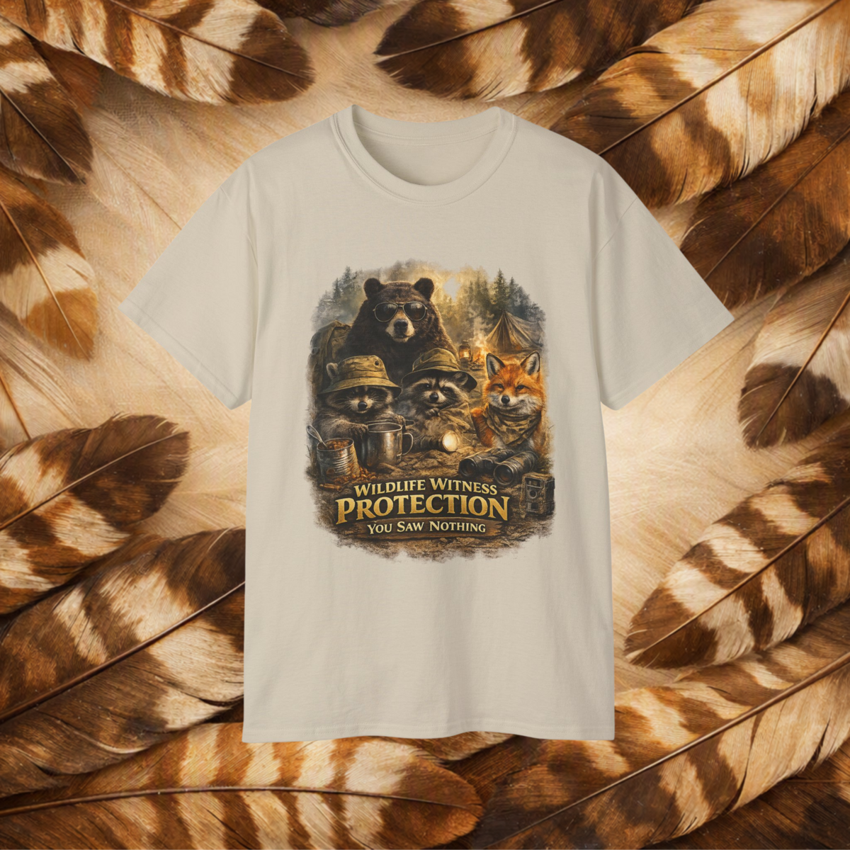 A Sand-colored short-sleeve t-shirt with a central graphic of raccoons, a bear, and a fox wearing hats or sunglasses, with the text 'WILDLIFE WITNESS PROTECTION YOU SAW NOTHING'. The shirt is displayed flat against a background of brown and beige feathers.