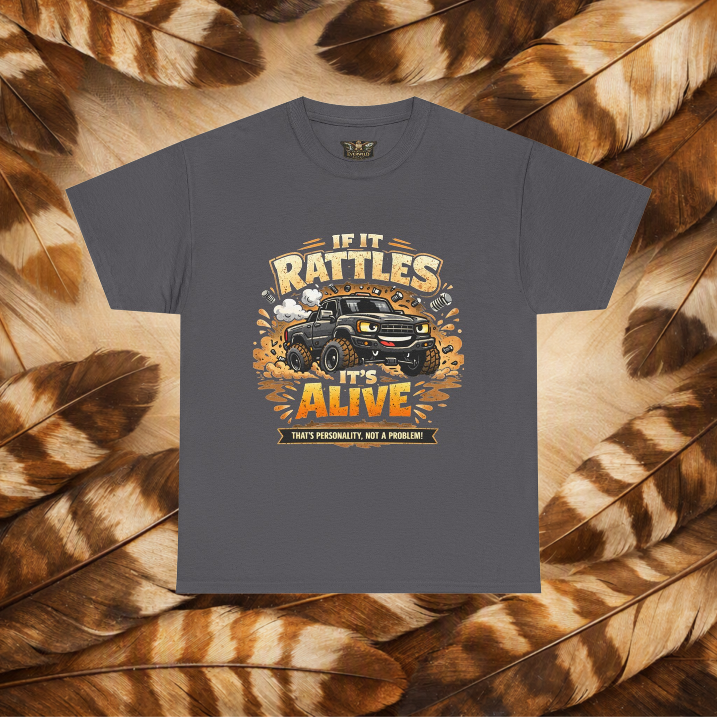 A black short-sleeve graphic t-shirt laid flat on a background of Hawk feathers. The graphic features a cartoon truck surrounded by text that reads 'IF IT RATTLES IT'S ALIVE' and 'THAT'S PERSONALITY, NOT A PROBLEM'.