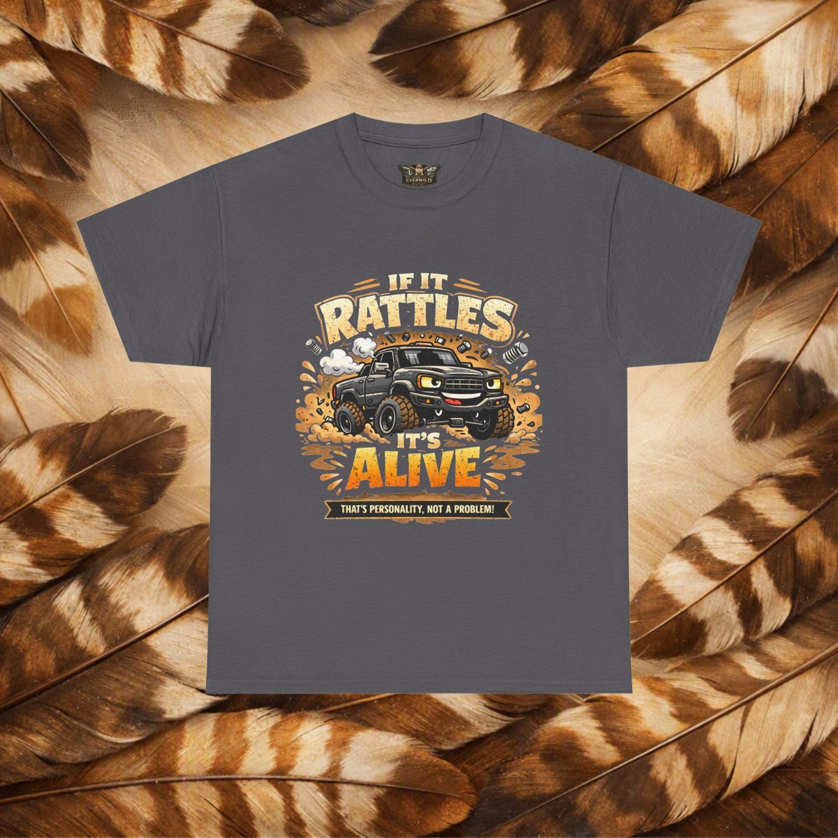 A black short-sleeve graphic t-shirt laid flat on a background of Hawk feathers. The graphic features a cartoon truck surrounded by text that reads 'IF IT RATTLES IT'S ALIVE' and 'THAT'S PERSONALITY, NOT A PROBLEM'.