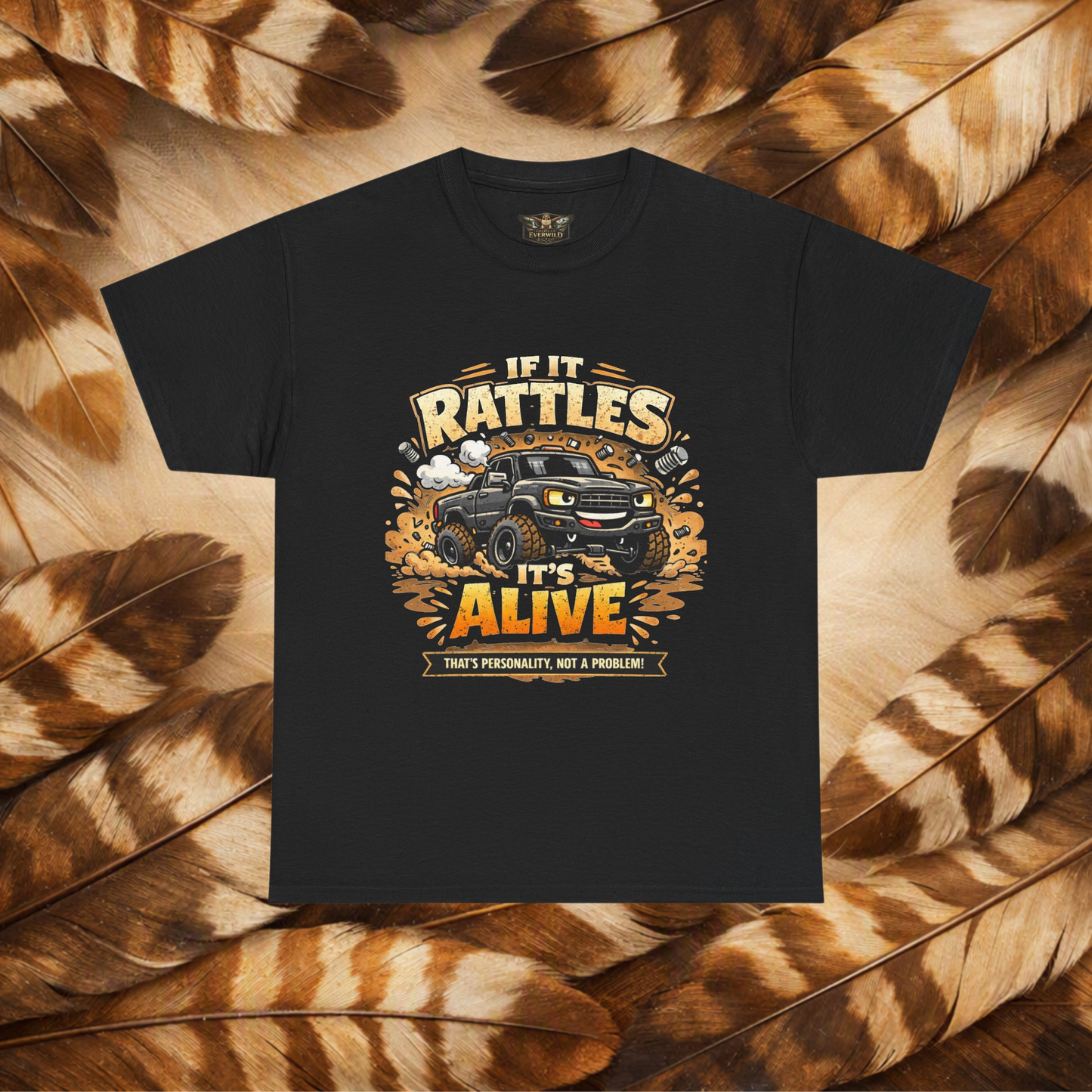 A black short-sleeve graphic t-shirt laid flat on a background of Hawk feathers. The graphic features a cartoon truck surrounded by text that reads 'IF IT RATTLES IT'S ALIVE' and 'THAT'S PERSONALITY, NOT A PROBLEM'.