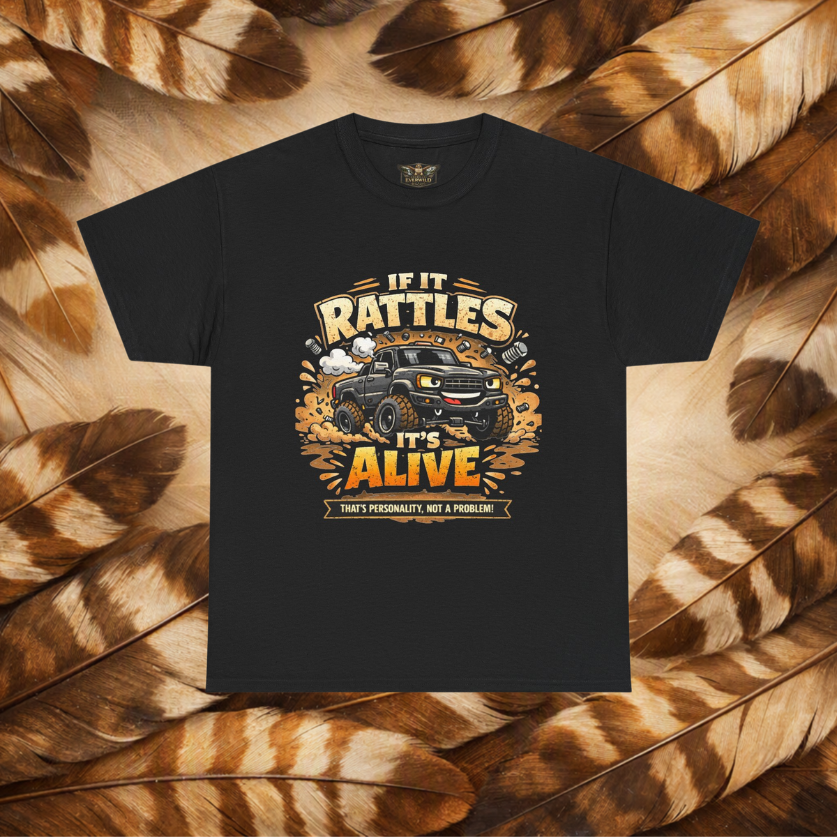 A black short-sleeve graphic t-shirt laid flat on a background of Hawk feathers. The graphic features a cartoon truck surrounded by text that reads 'IF IT RATTLES IT'S ALIVE' and 'THAT'S PERSONALITY, NOT A PROBLEM'.