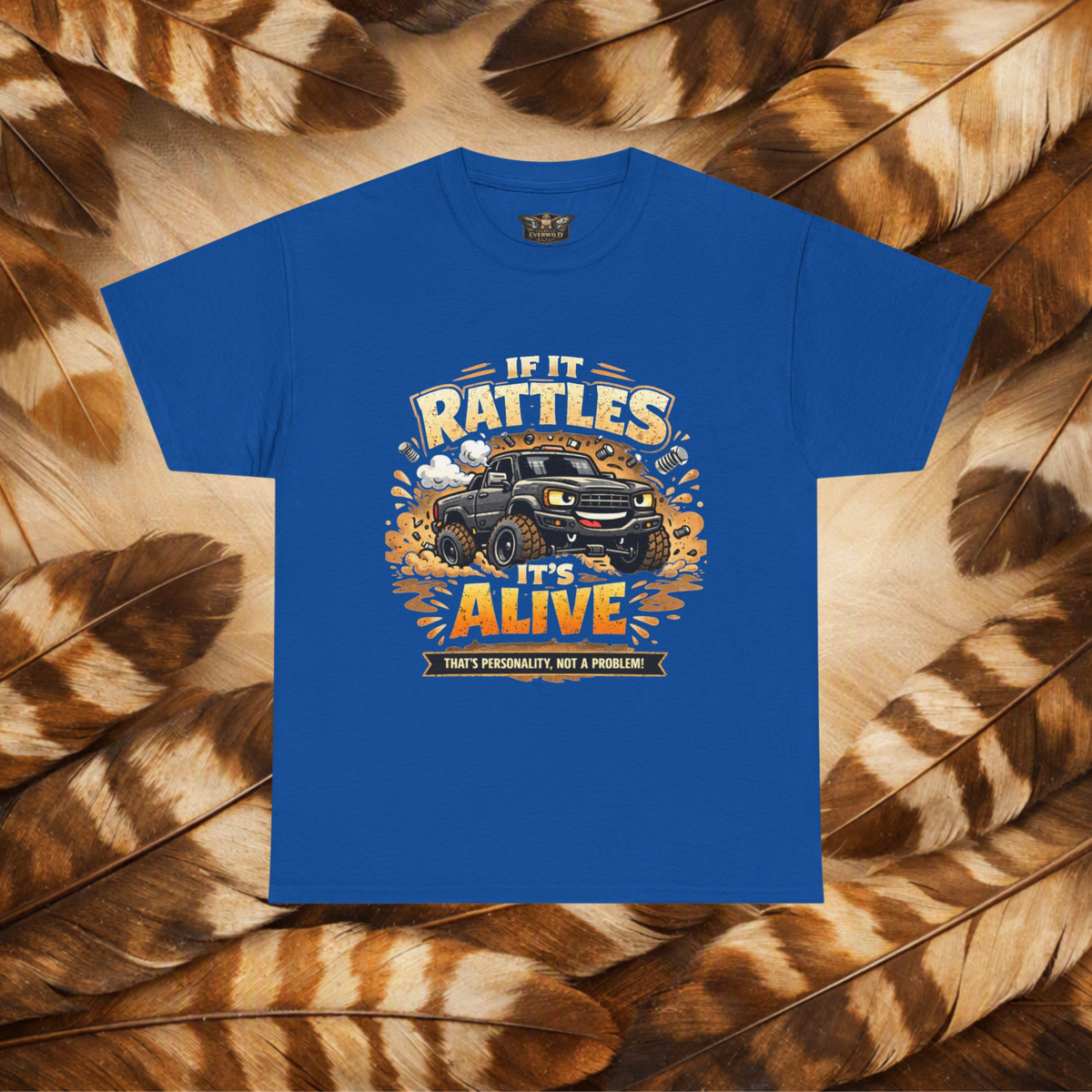 A Royal Blue_short-sleeve graphic t-shirt laid flat on a background of Hawk feathers. The graphic features a cartoon truck surrounded by text that reads 'IF IT RATTLES IT'S ALIVE' and 'THAT'S PERSONALITY, NOT A PROBLEM'.