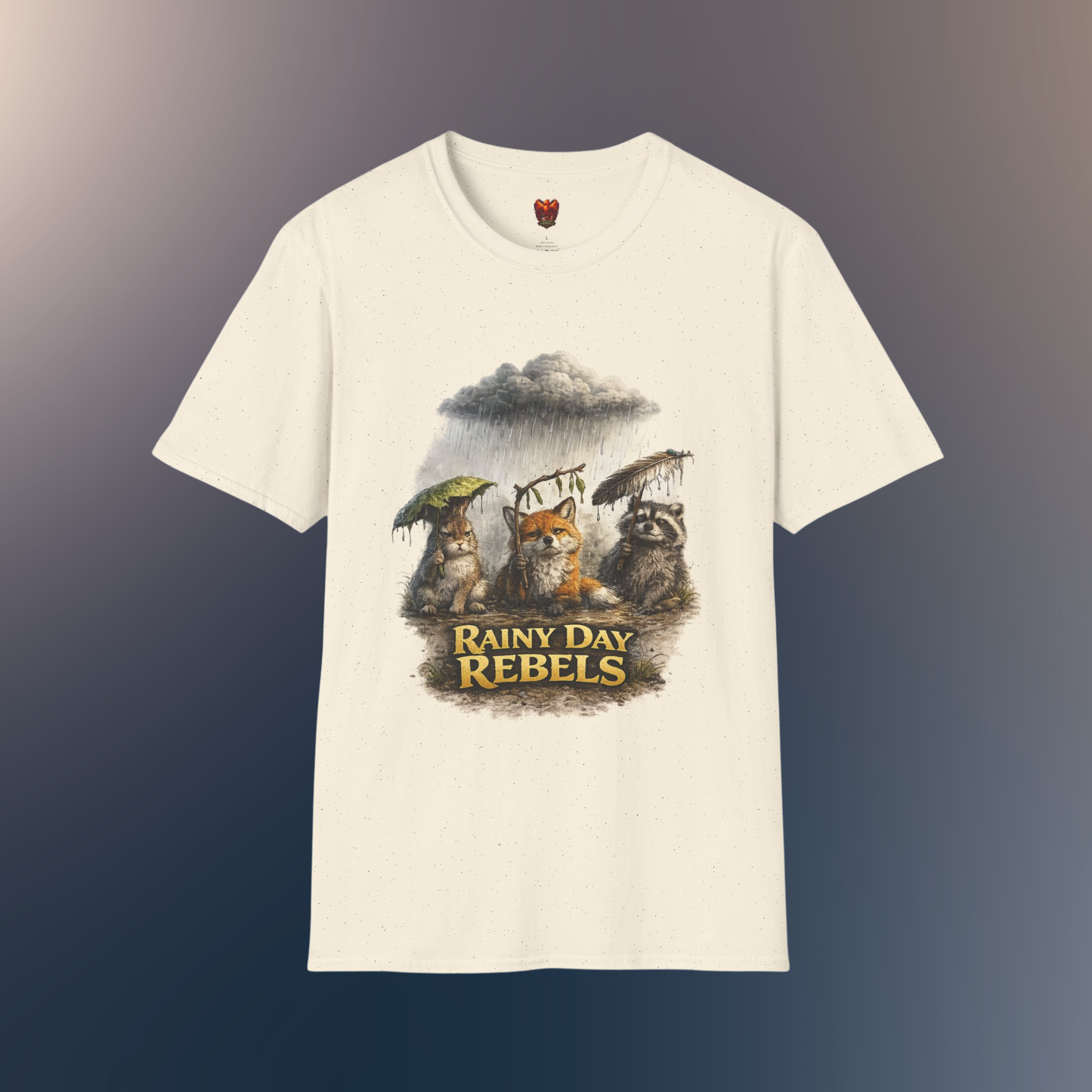 An natural short-sleeve crewneck t-shirt featuring a graphic print of three forest animals holding umbrellas under a rainy storm cloud, with the text 'RAINY DAY REBELS' below them, on a hawk feather flat lay background.