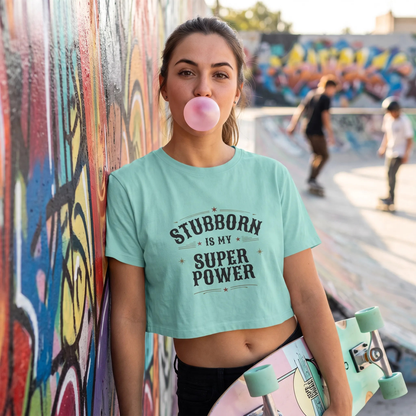 A woman in a skatepark wearing a chalky mint crop top with graphic that reads 'STUBORN IS MY SUPER POWER'. She is holding a skateboard and blowing a pink bubblegum bubble against a colorful graffiti wall.