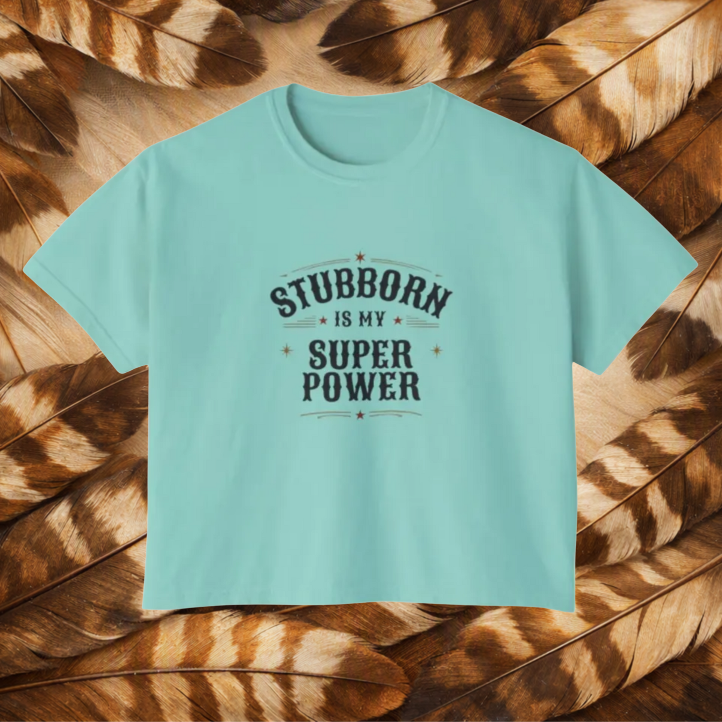 A chalky mint color short-sleeve t-shirt with a dark gray graphic text reading 'STUBBORN IS MY SUPER POWER' laid flat on a background of brown and tan feathers.