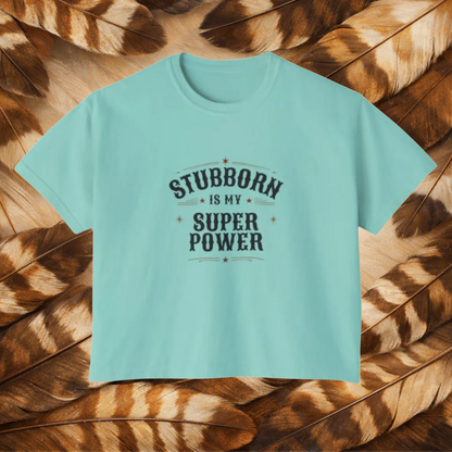 A chalky mint color short-sleeve t-shirt with a dark gray graphic text reading 'STUBBORN IS MY SUPER POWER' laid flat on a background of brown and tan feathers.