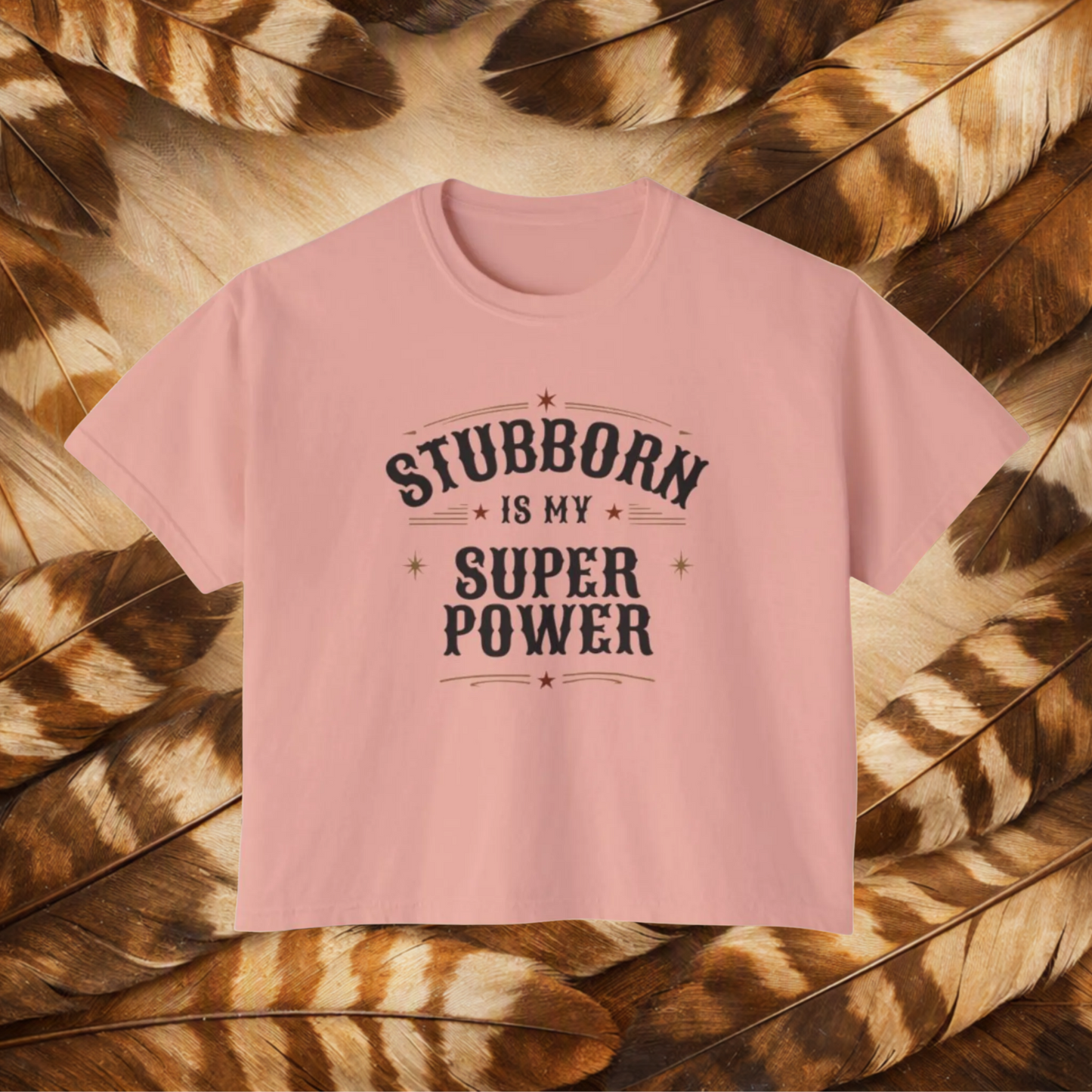 A peachy color short-sleeve t-shirt with a dark gray graphic text reading 'STUBBORN IS MY SUPER POWER' laid flat on a background of brown and tan feathers.