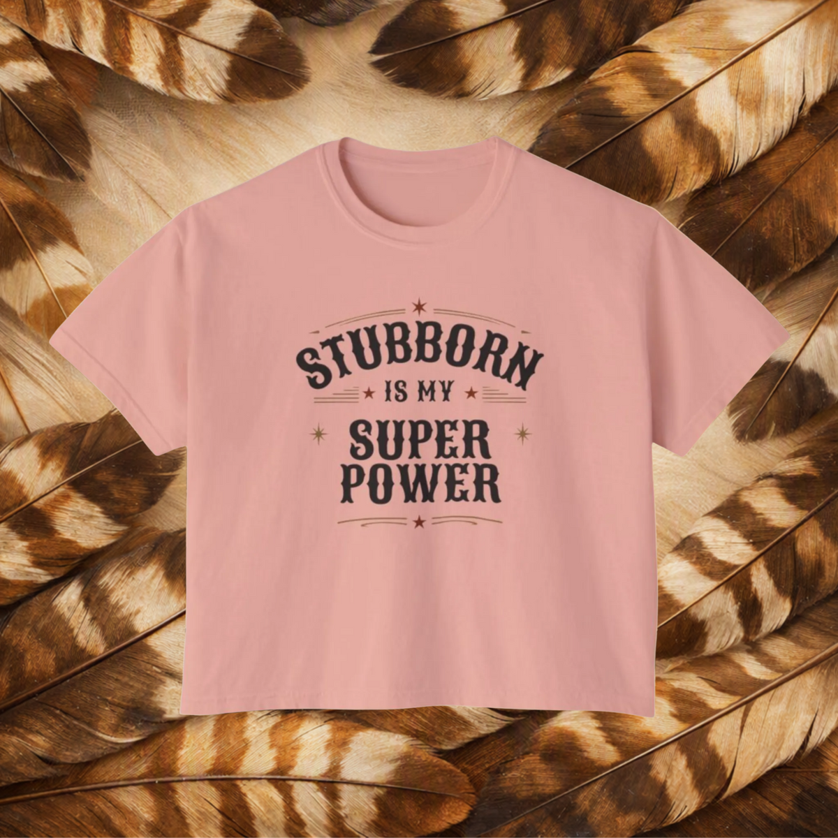 A peachy color short-sleeve t-shirt with a dark gray graphic text reading 'STUBBORN IS MY SUPER POWER' laid flat on a background of brown and tan feathers.