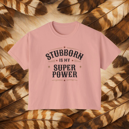 A peachy color short-sleeve t-shirt with a dark gray graphic text reading 'STUBBORN IS MY SUPER POWER' laid flat on a background of brown and tan feathers.