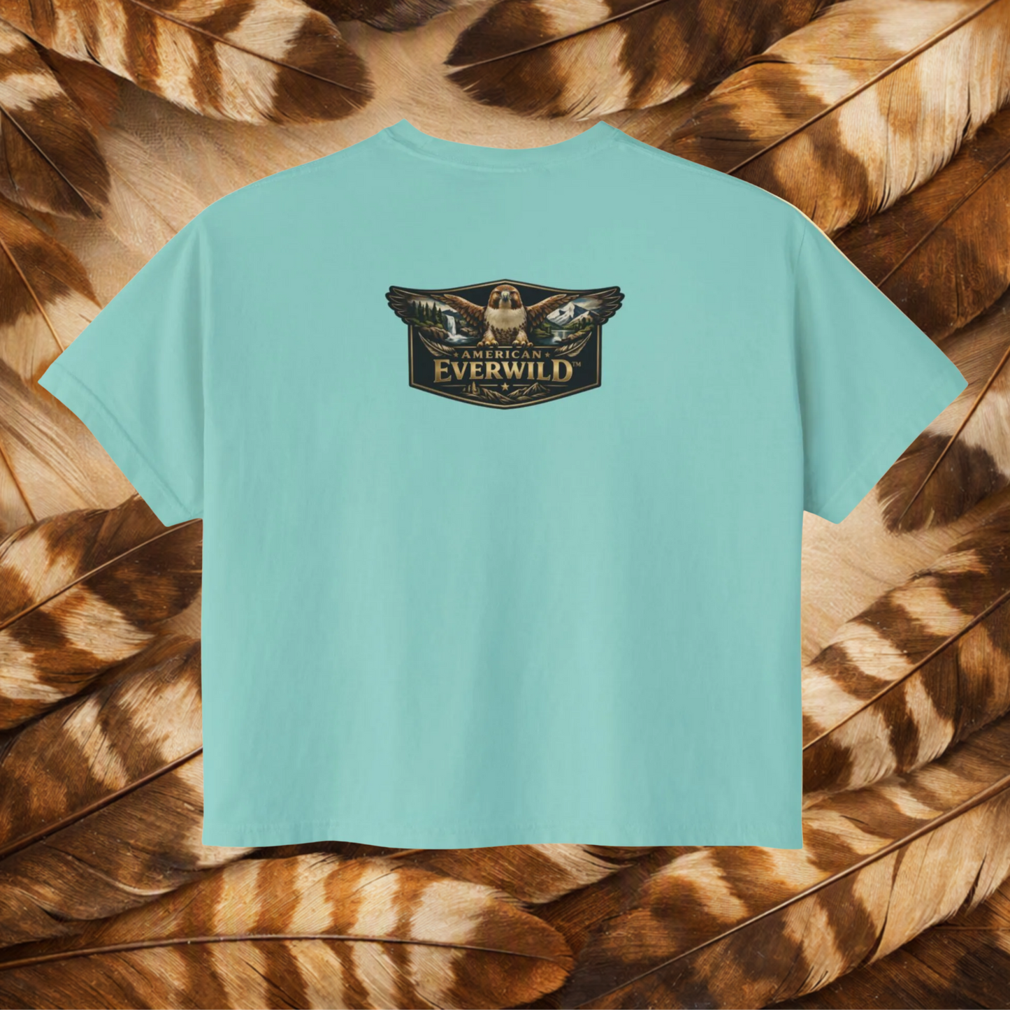 A chalky mint short-sleeve t-shirt with a central graphic of a hawk with wings outstreched with painted scene in the wings and the text 'American Everwild', displayed flat against a background of brown and tan feathers.