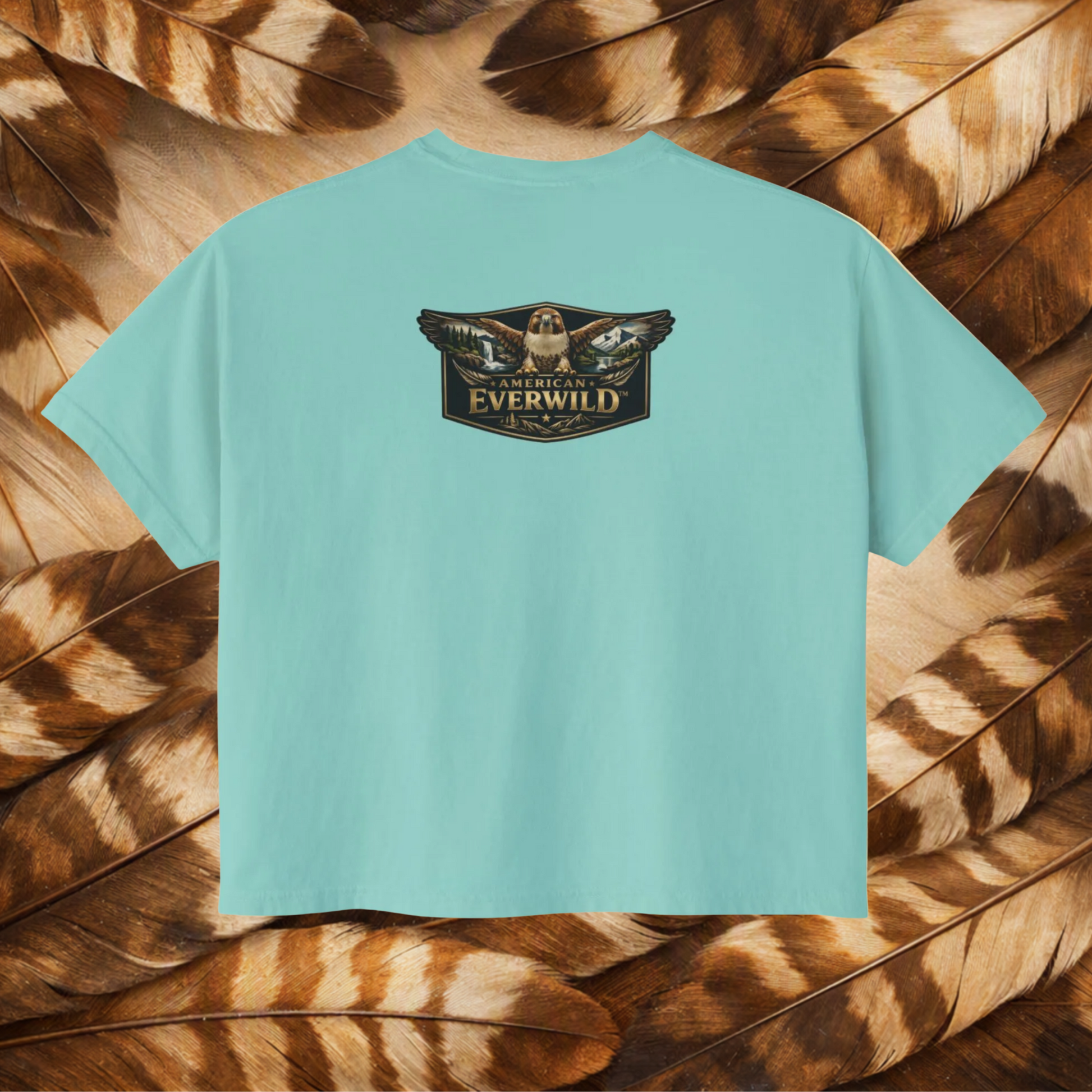 A chalky mint short-sleeve t-shirt with a central graphic of a hawk with wings outstreched with painted scene in the wings and the text 'American Everwild', displayed flat against a background of brown and tan feathers.