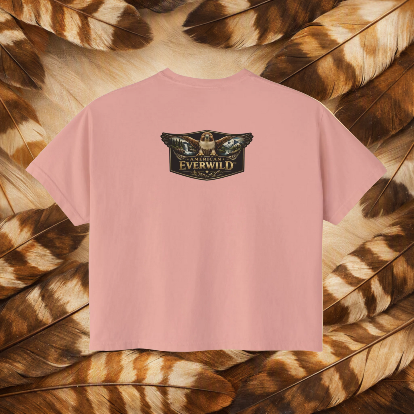 A peachy short-sleeve t-shirt with a central graphic of a hawk with wings outstreched with painted scene in the wings and the text 'American Everwild', displayed flat against a background of brown and tan feathers.